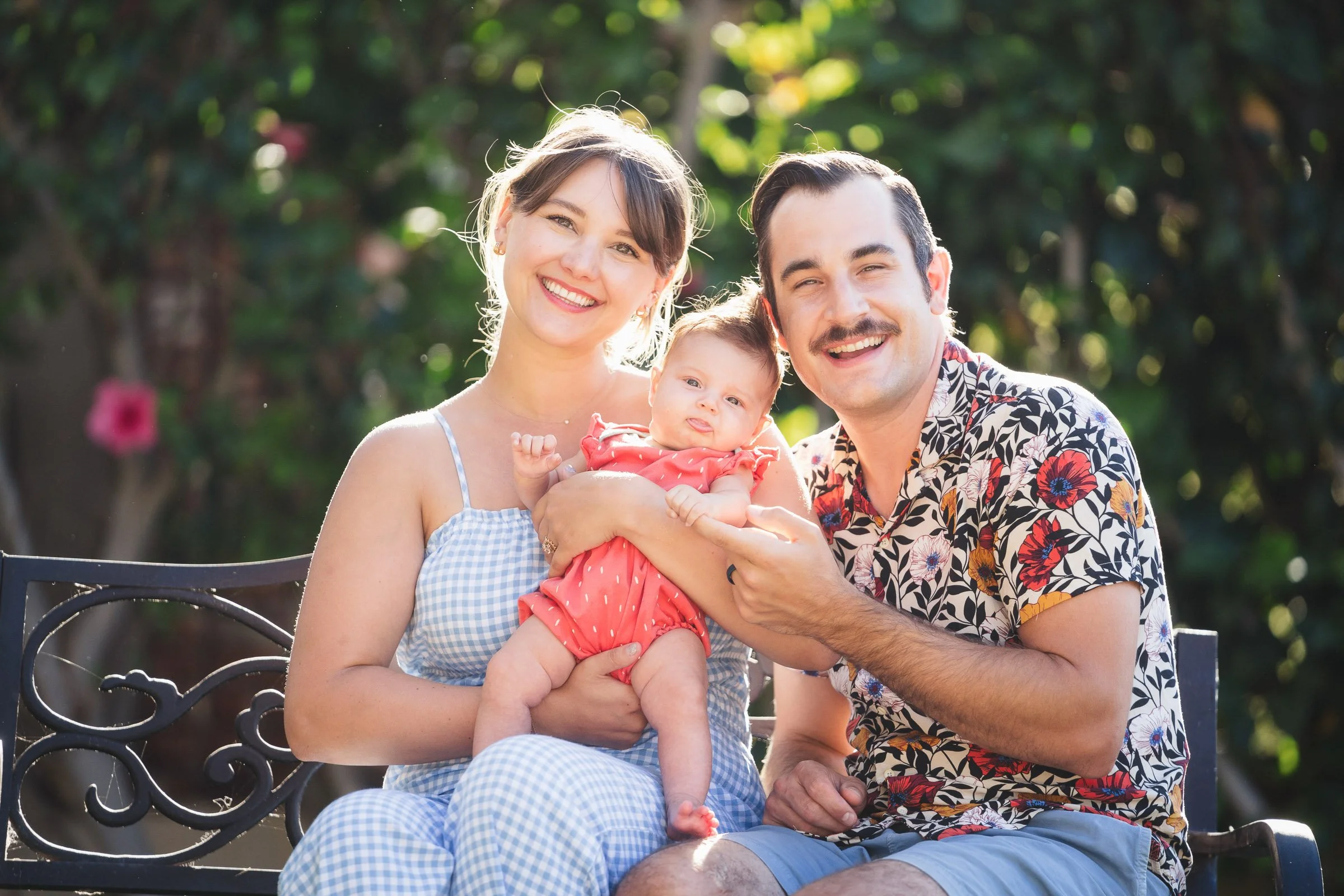 Smiling man and woman sitting on a bench outdoors, holding a baby girl with light skin and dark hair, with a background of green trees and sunlight.