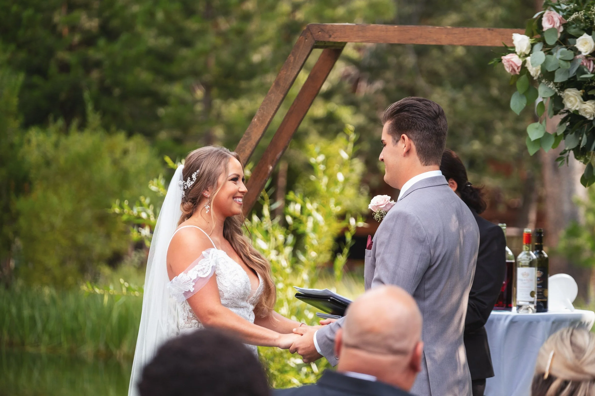 Bride and groom holding hands and smiling during an outdoor wedding ceremony in a forested area, with a wedding officiant and guests in the background.
