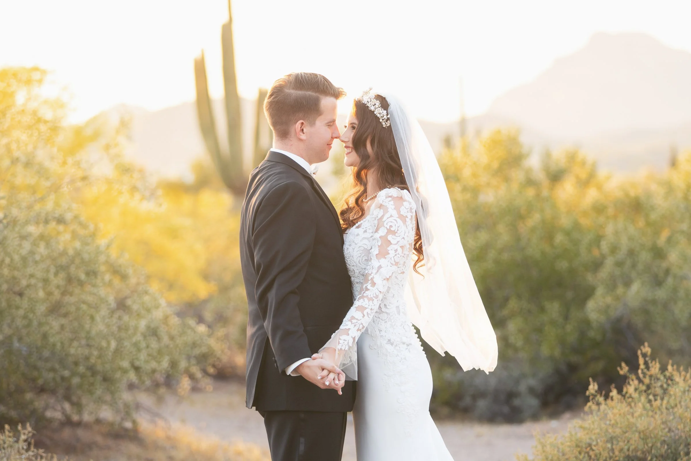 A newlywed couple in wedding attire holding hands in a desert landscape with cacti and mountains in the background, during sunset.
