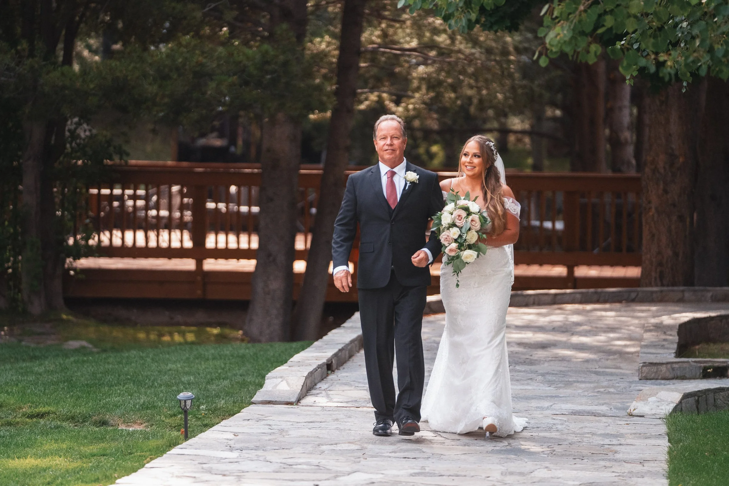 A bride in a white wedding gown holding a bouquet of roses walks arm in arm with a man in a dark suit, possibly her father, outdoors on a stone pathway lined with grass, surrounded by trees.