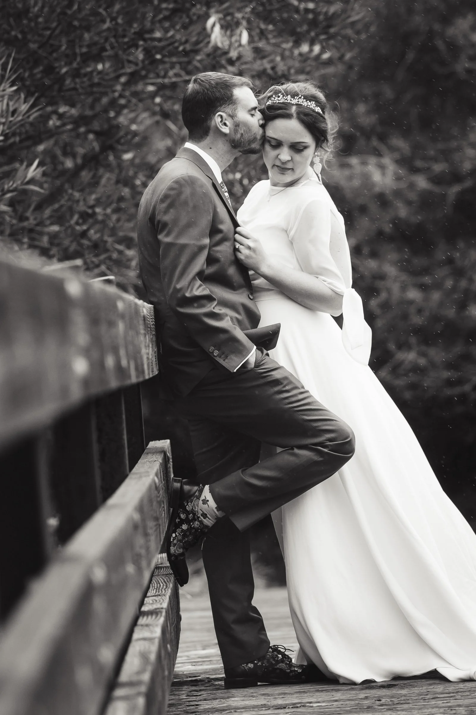 Black and white photo of a bride and groom; the groom is kissing the bride's forehead while she looks down, sitting on a wooden bridge with a blurry natural background.