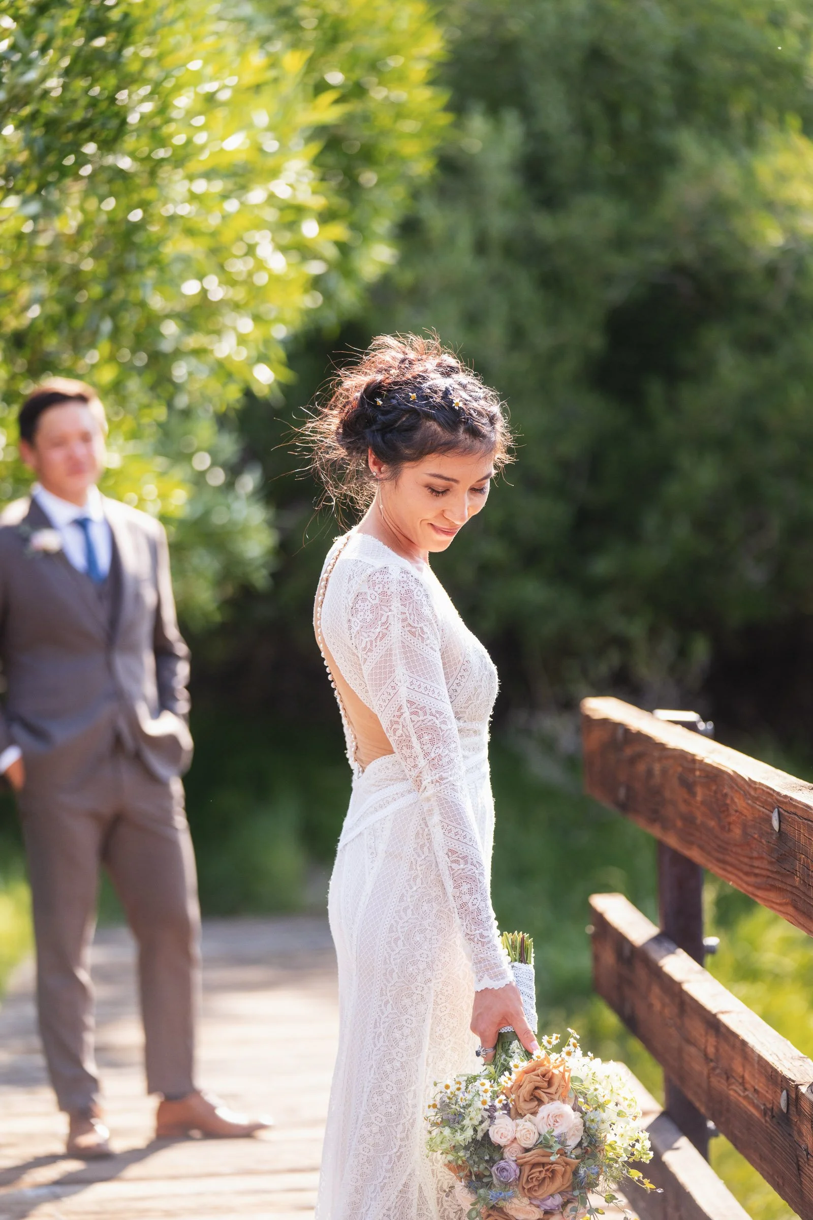 A bride in a white lace wedding dress holding a bouquet of flowers, standing on a wooden bridge outdoors with greenery, while a groom in a suit stands in the background.
