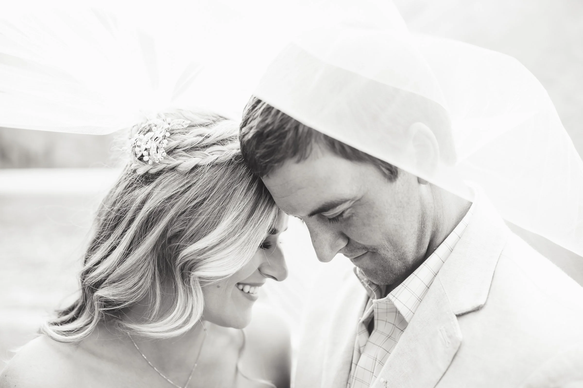 A black and white photo of a smiling woman and a man with their foreheads touching, sharing an intimate moment in wedding attire.