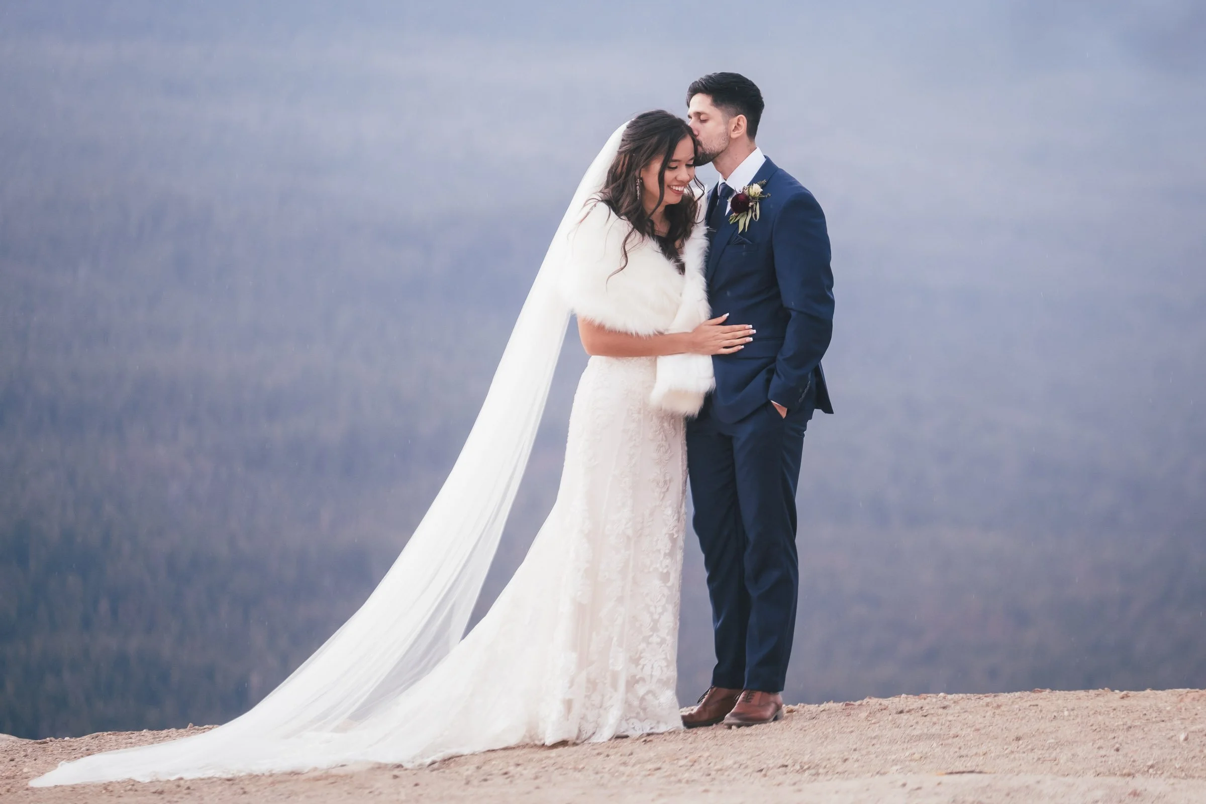 A bride and groom sharing an intimate moment on a rocky outdoor terrain with mountains in the background. The bride is wearing a white wedding dress with a long veil and fur shawl, and the groom is dressed in a navy blue suit with a boutonniere.