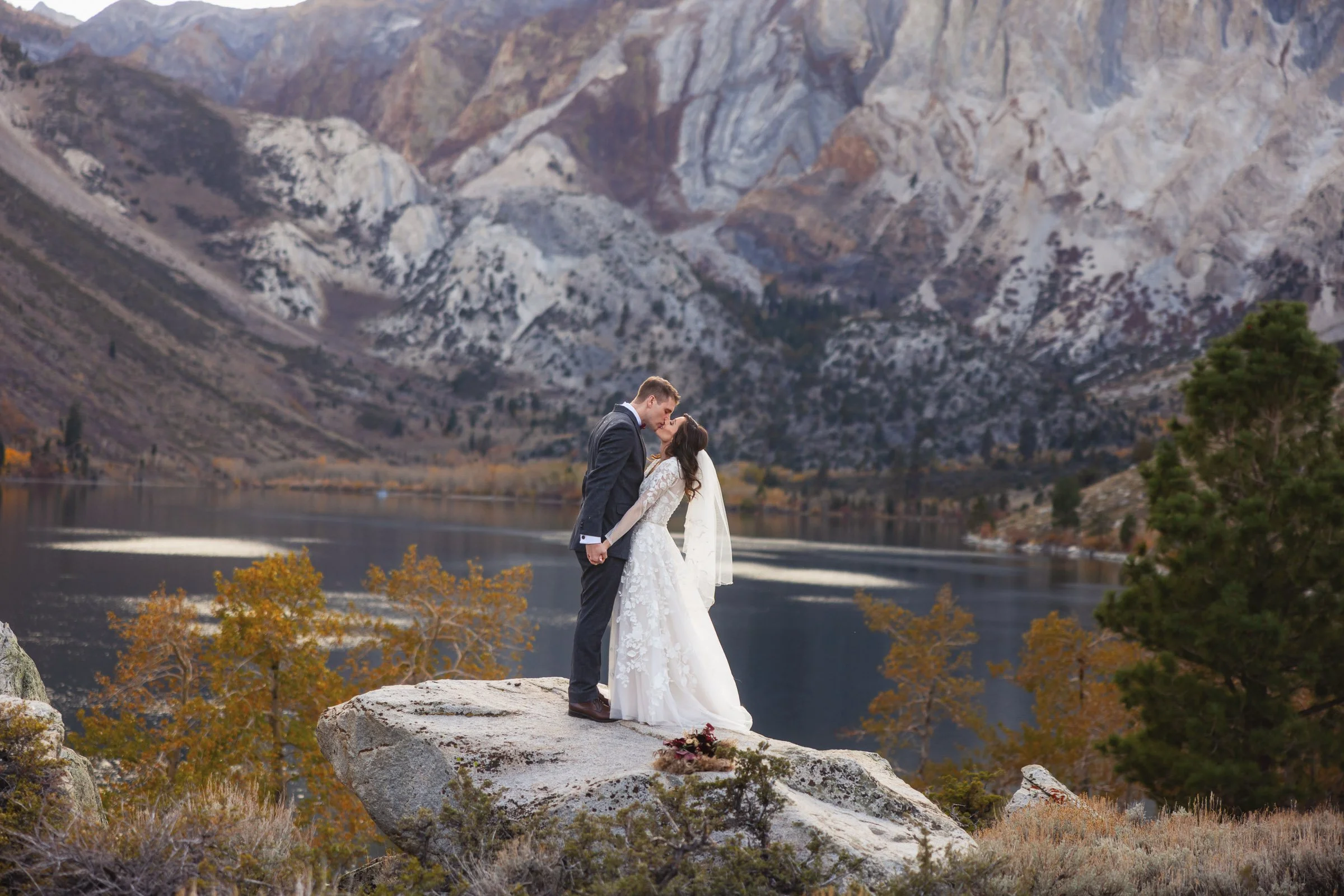 A couple in wedding attire sharing a kiss on a large rock in a mountainous outdoor setting with a lake and autumn trees in the background.