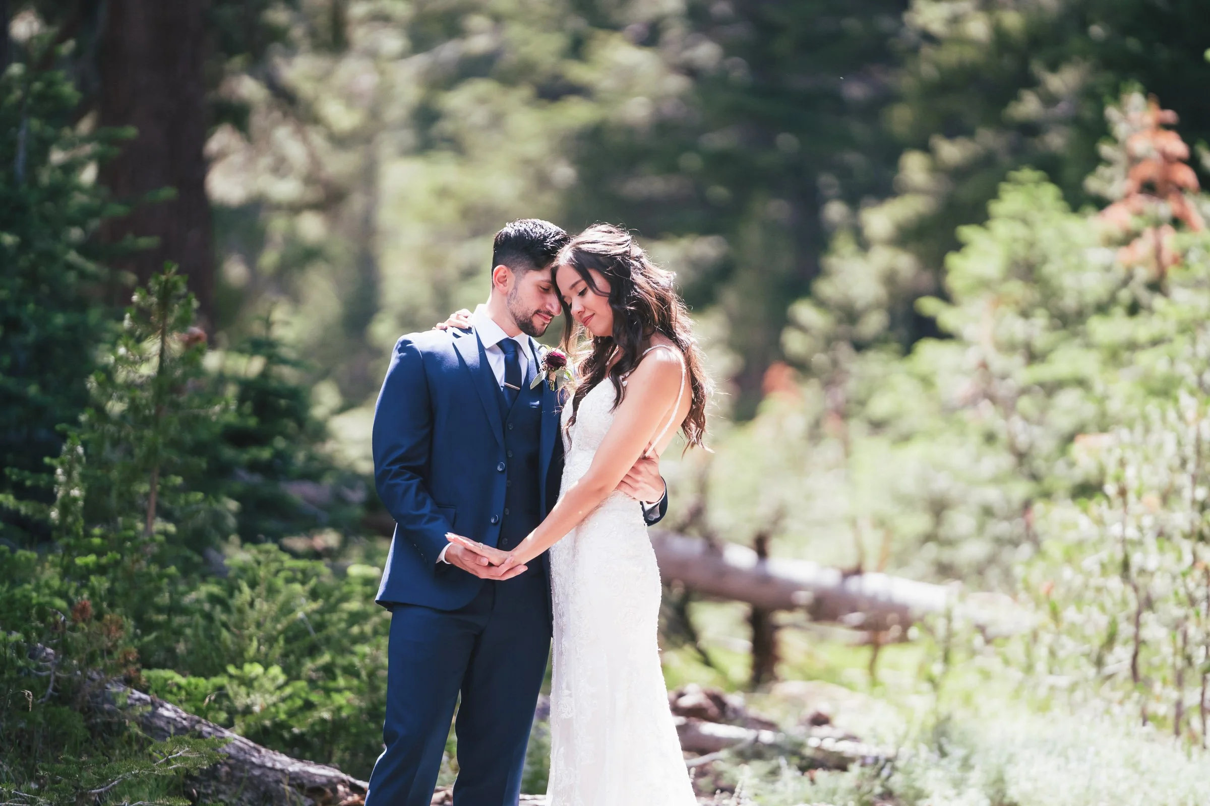 A newlywed couple in wedding attire standing close together, holding hands, in a lush green forest setting.