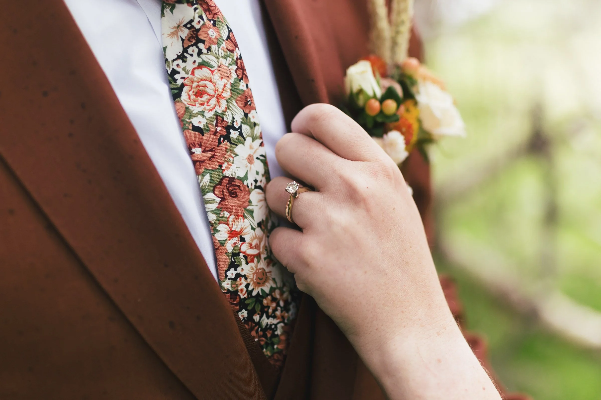 Close-up of a woman wearing a suit with a floral patterned tie, holding her hand near her chest with an engagement ring visible on her finger, and a mixed flower boutonniere pinned to her suit.