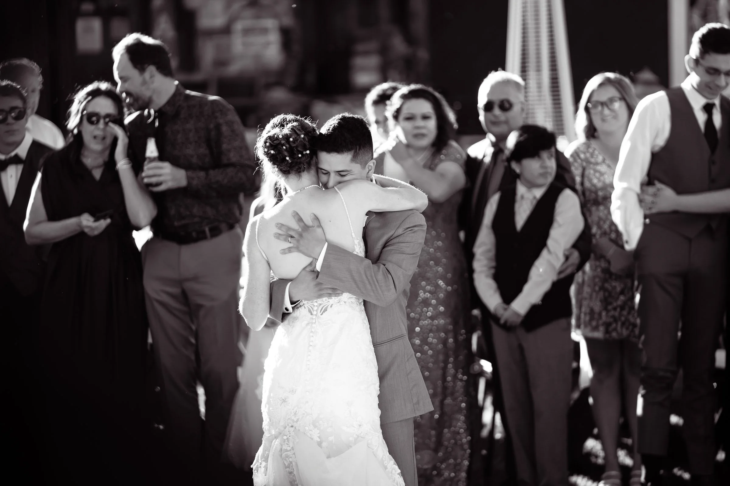 A wedding couple shares a hug as guests look on, in black and white.