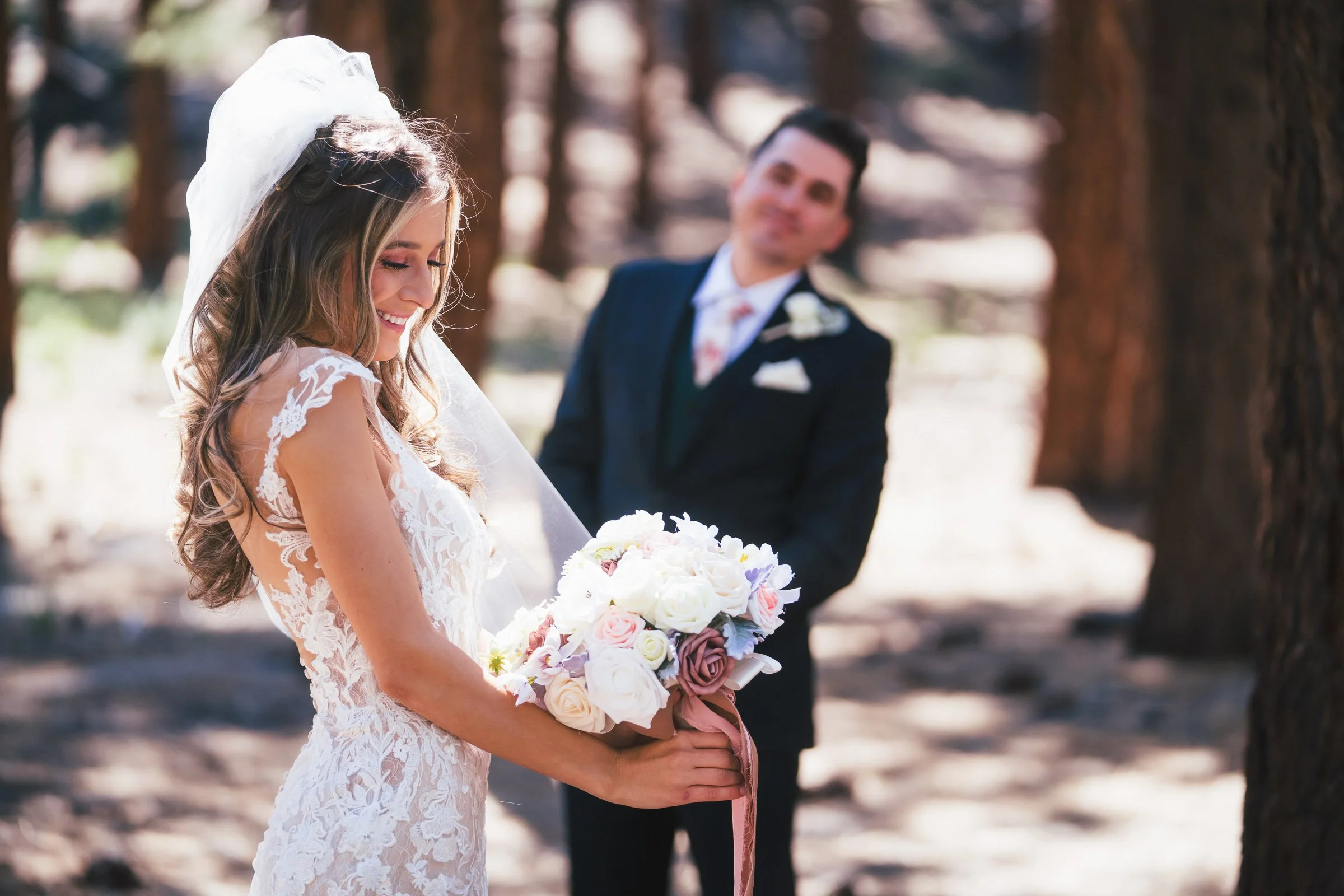 A bride holding a bouquet of roses smiles as she looks down, while a groom in a dark suit smiles in the background, standing among trees in a forest setting.