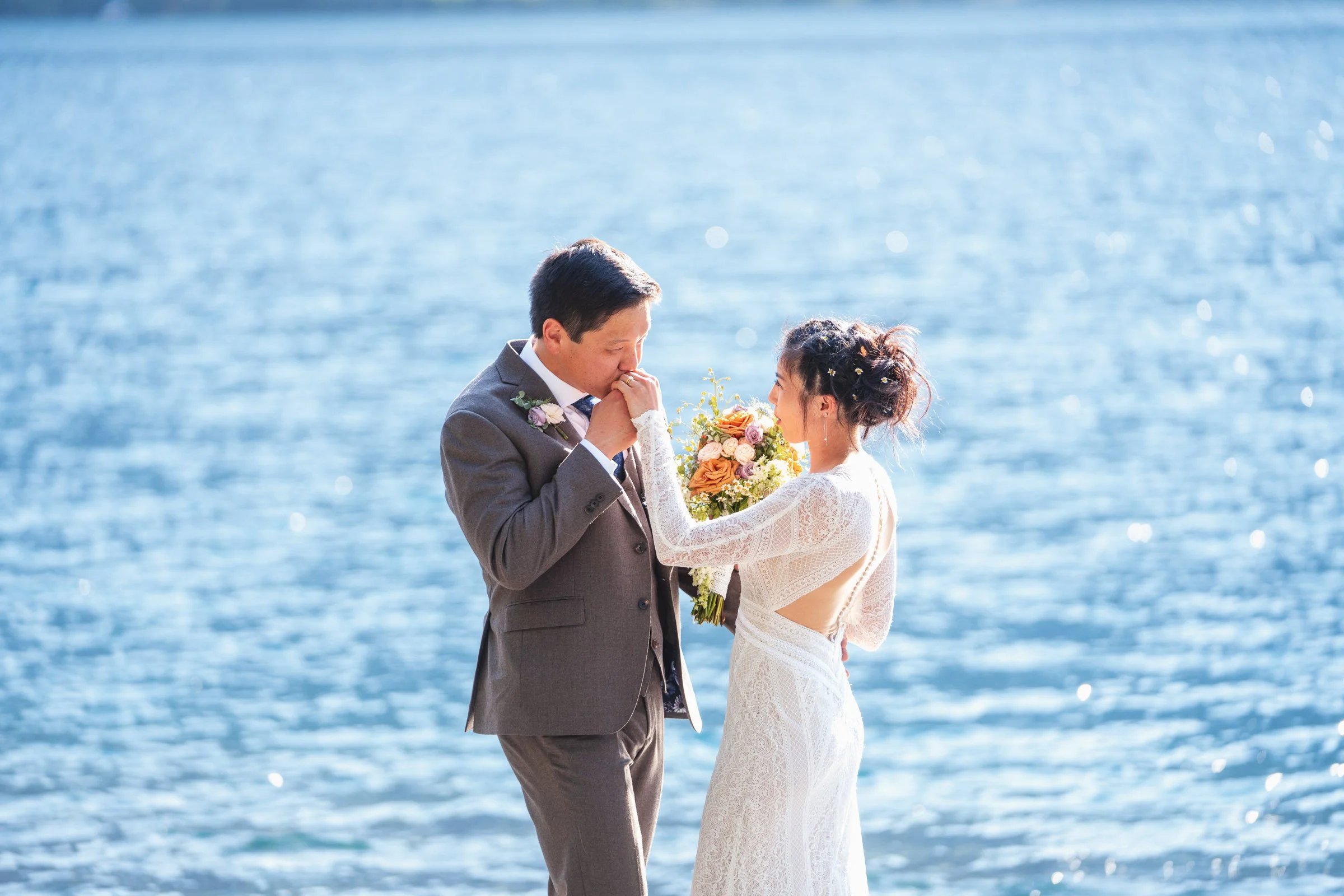 A bride and groom stand at a waterfront, sharing an intimate moment. The groom is gently holding the bride's hand to his lips, while the bride holds a bouquet of flowers. The background features a sparkling blue body of water.
