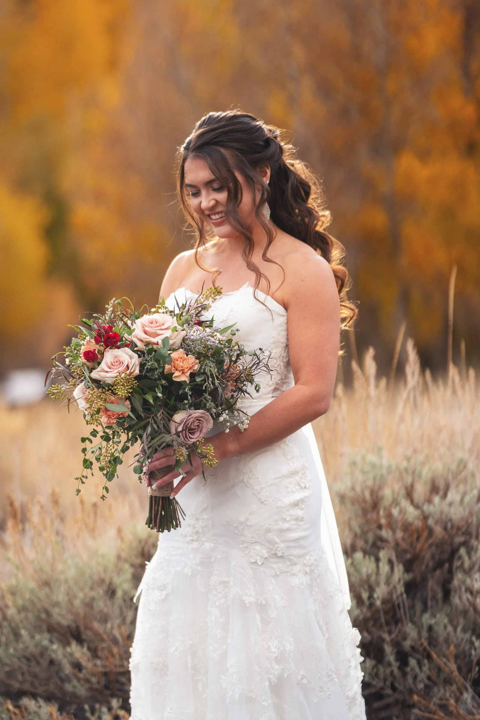 Bride in a white wedding dress holding a bouquet of pink and red flowers with green foliage, smiling outdoors with autumn foliage in the background.