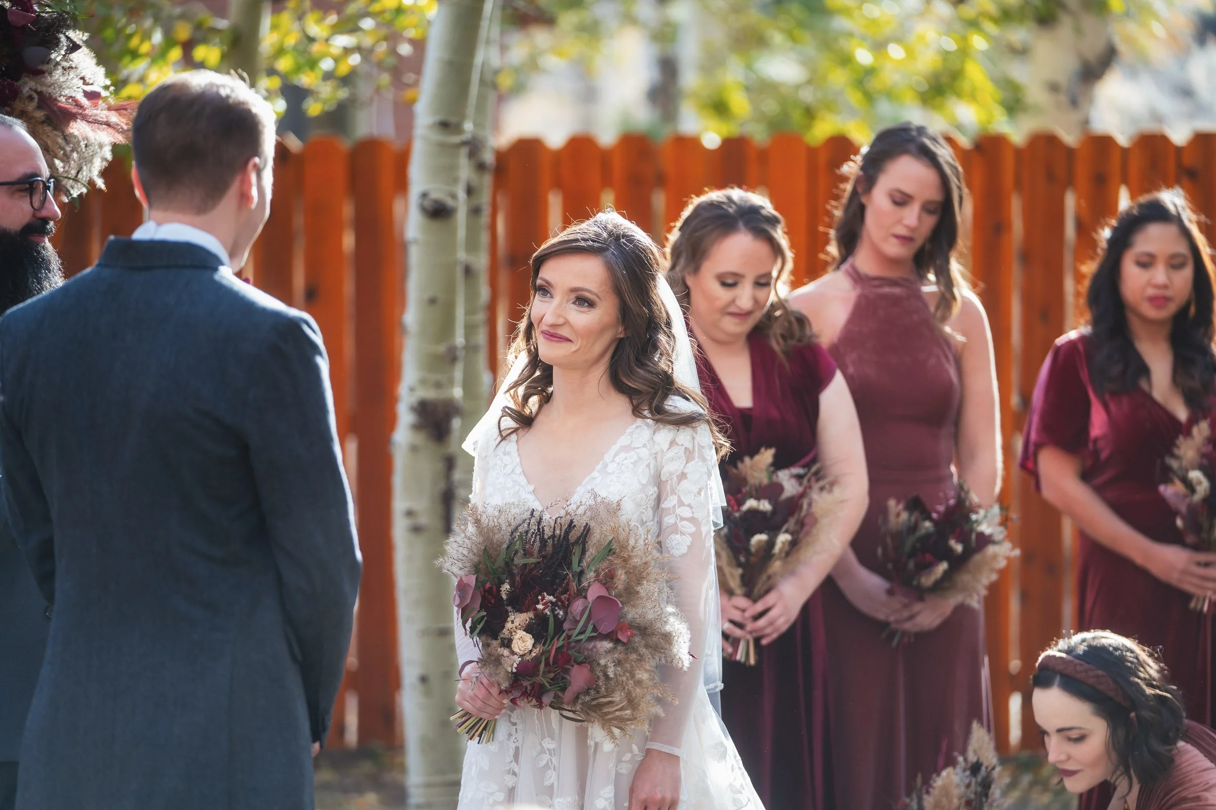 A woman in a white wedding dress holding a bouquet looks at a man in a gray suit during an outdoor wedding ceremony, with bridesmaids in burgundy dresses holding bouquets in the background.