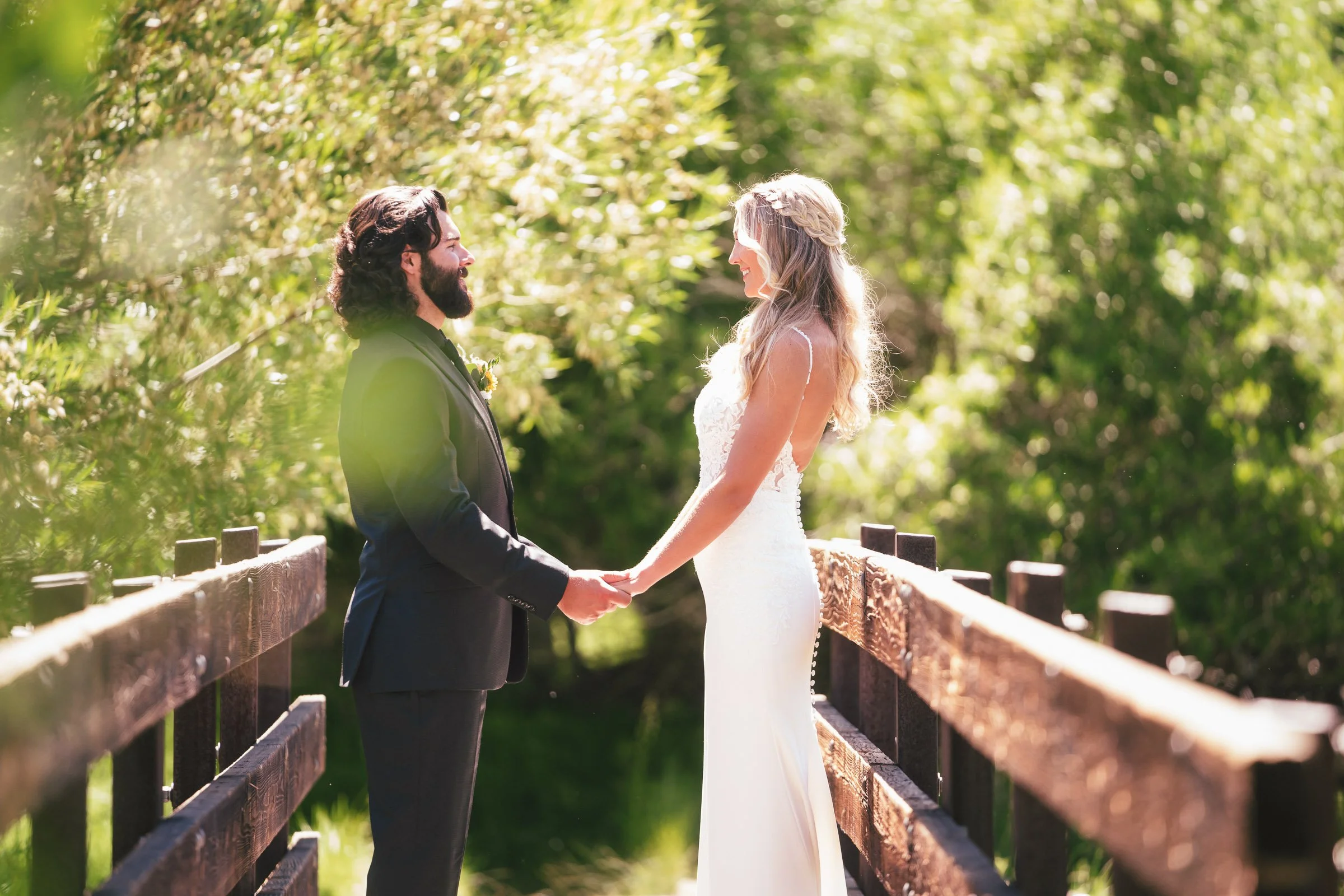 A bride and groom standing on a wooden bridge, holding hands and facing each other in a sunny, lush green outdoor setting.
