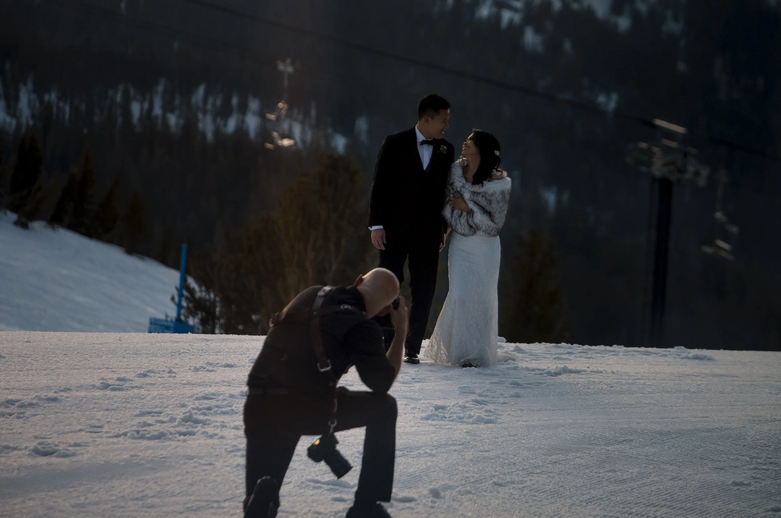 A bride and groom in wedding attire holding each other and smiling on a snowy hilltop during dusk, with a photographer kneeling in front capturing the moment.