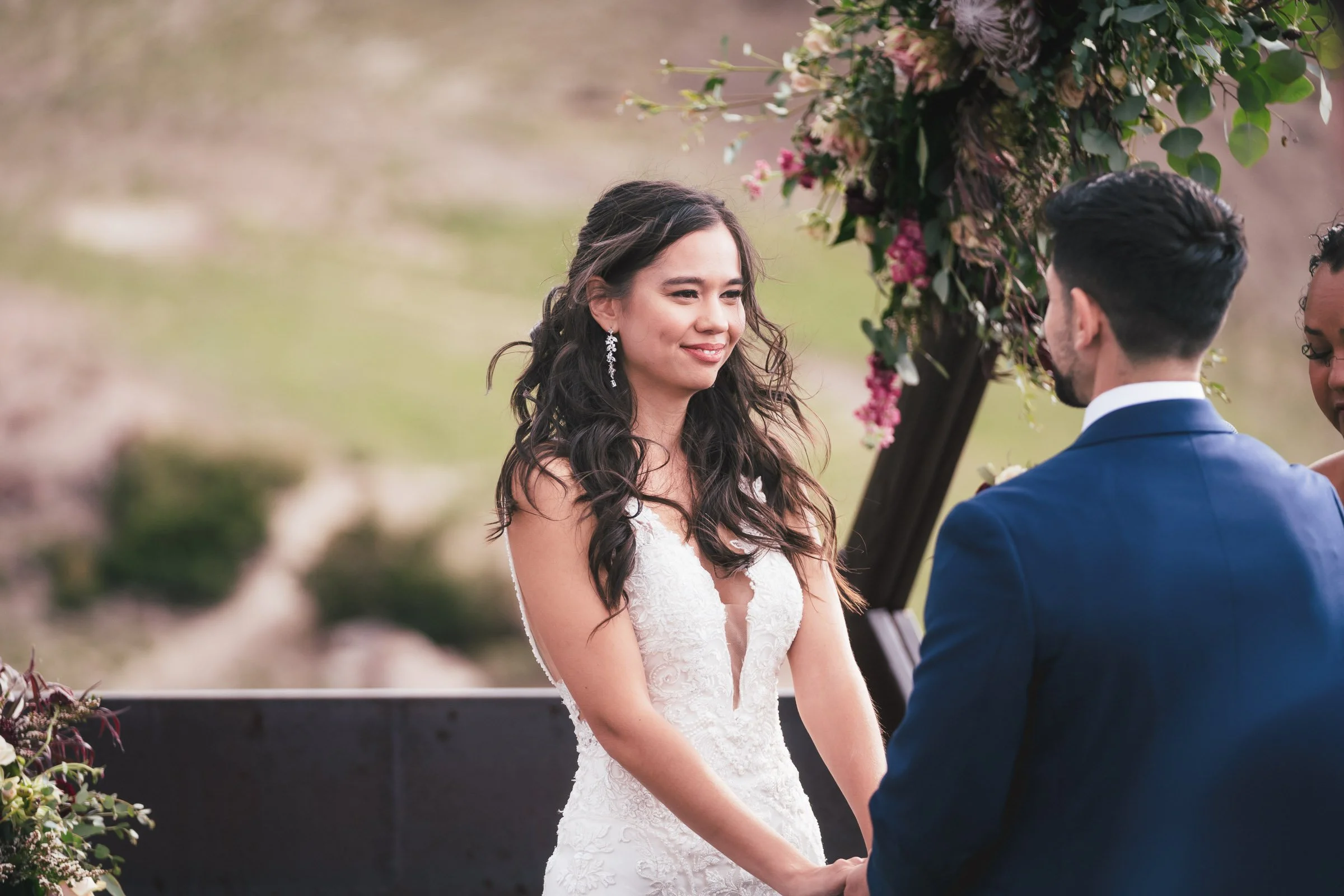 A woman in a white wedding dress and long earrings holds hands with a man in a navy suit during their outdoor wedding ceremony, standing under a floral arch with a blurred natural background.