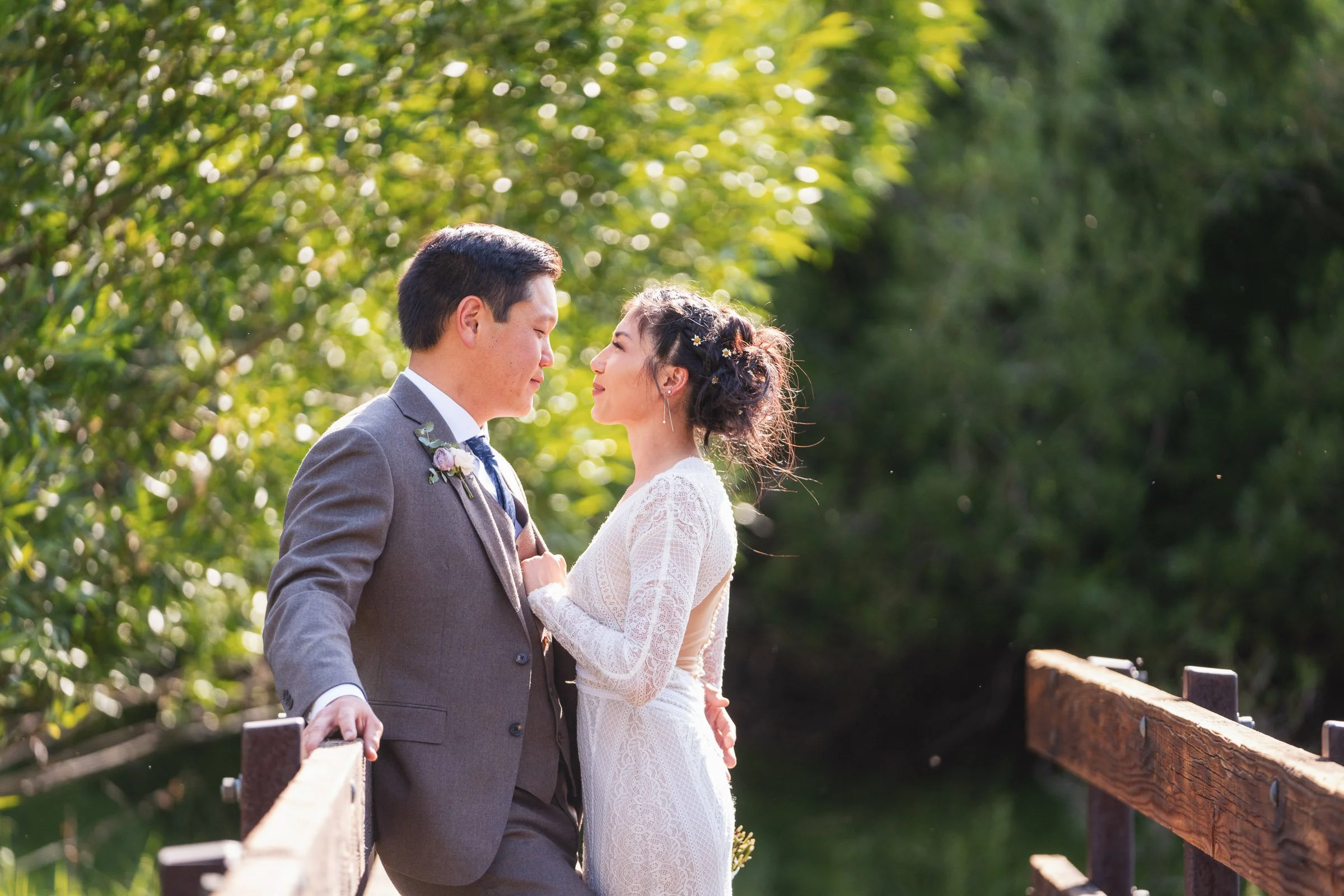 A bride and groom standing close on a wooden bridge, facing each other, outdoors surrounded by green trees, during daytime.