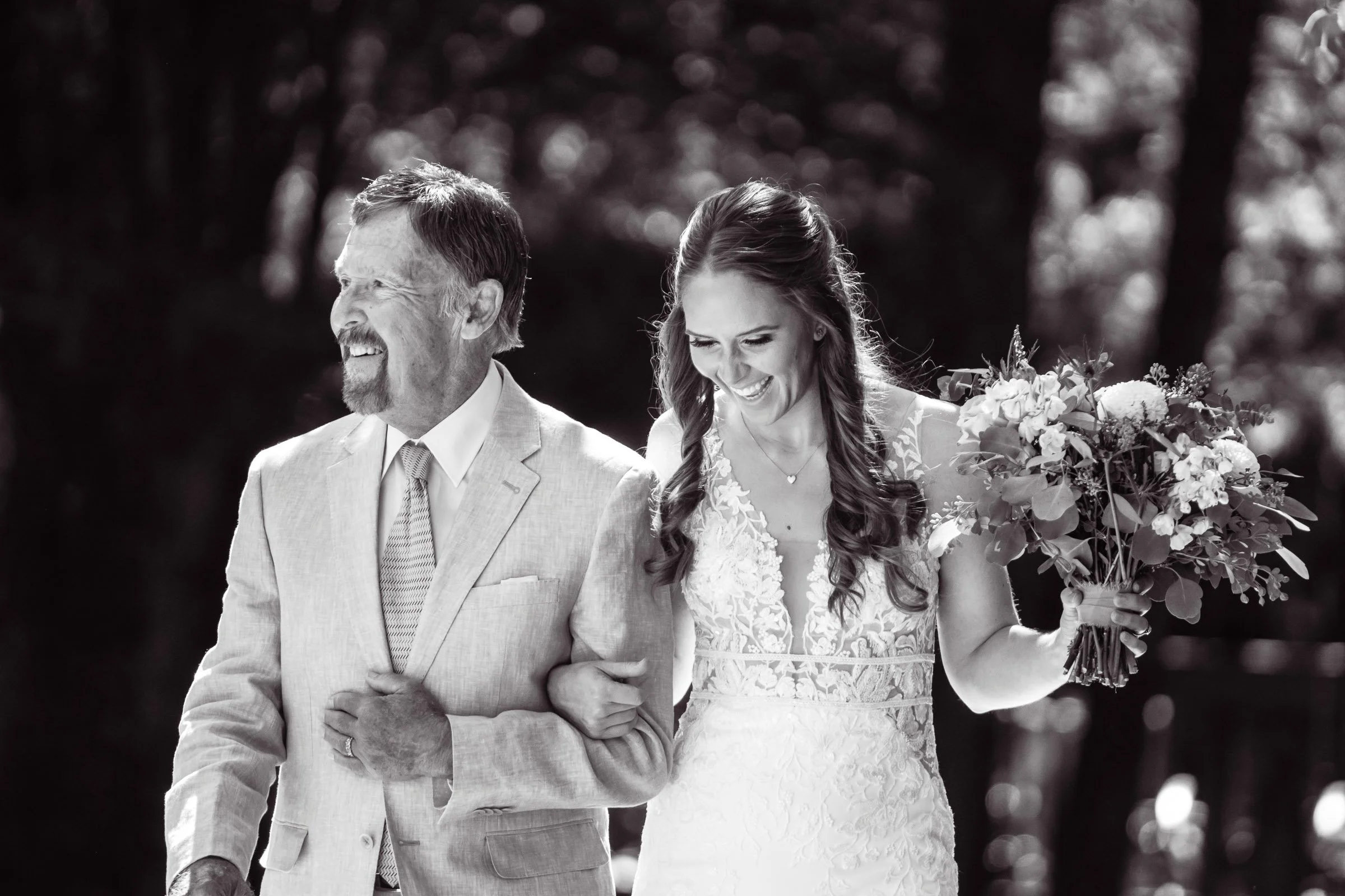 A bride smiling and holding a bouquet of flowers in her right hand, walking arm-in-arm with an older man, outdoors, black and white photograph.
