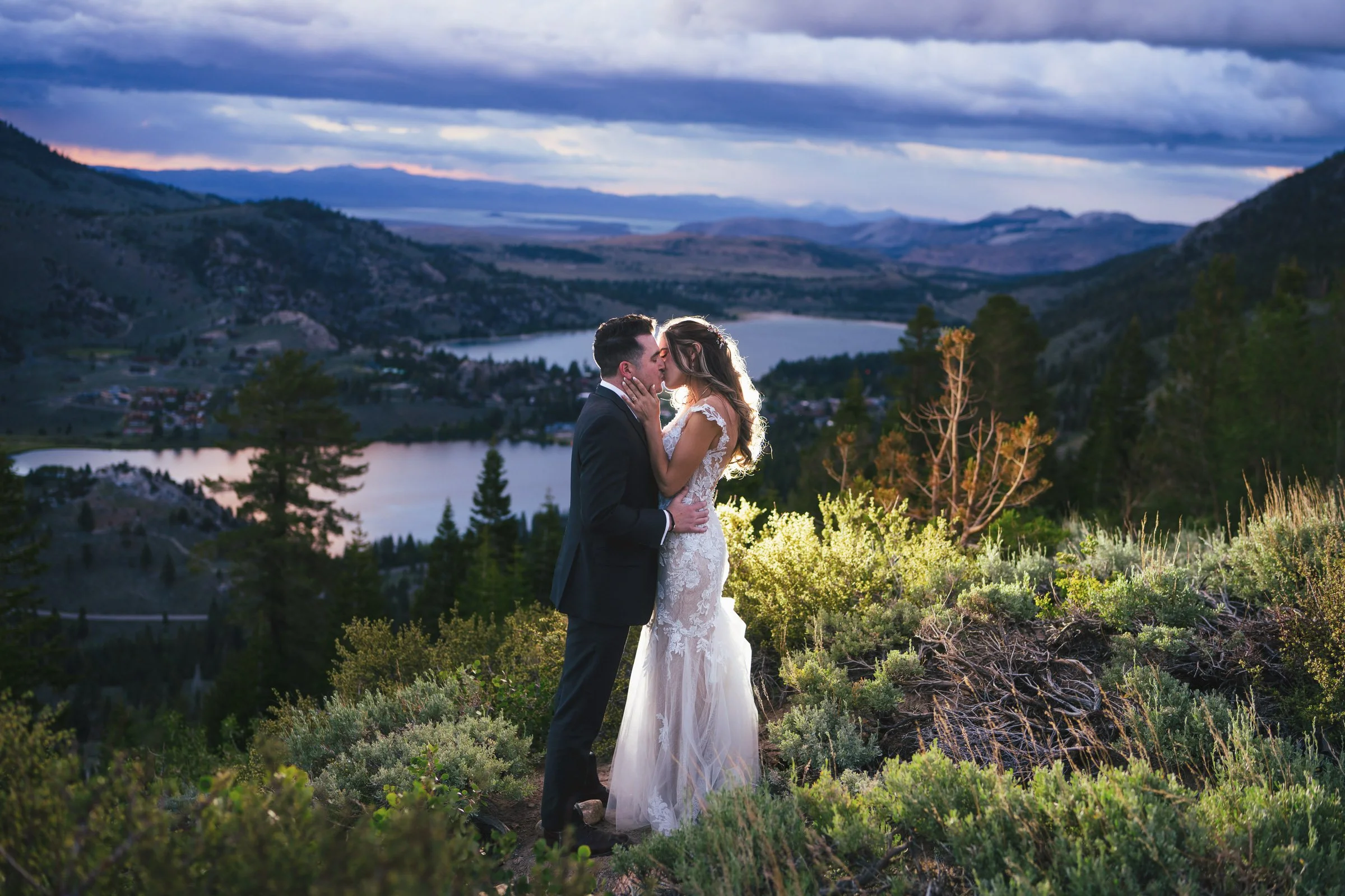 A newlywed couple kissing in a scenic outdoor setting with a lake and mountainous landscape at dusk.