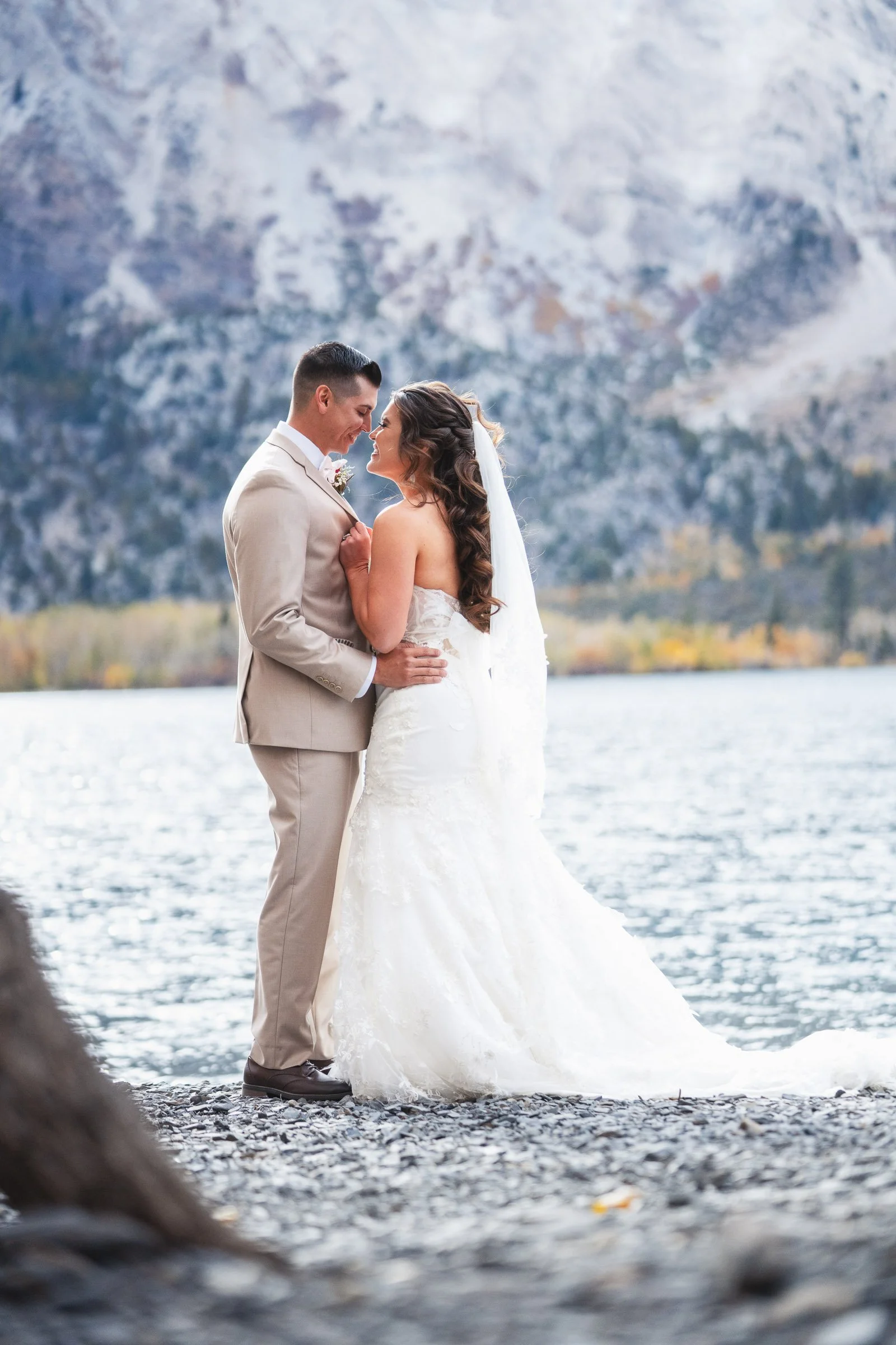 A bride and groom standing close by a lake with mountain scenery in the background, sharing a moment on their wedding day.