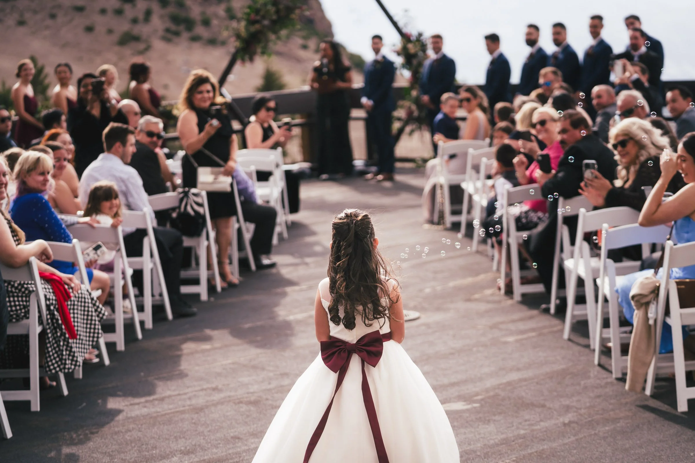 A young girl in a white dress with a large maroon bow in the back stands facing an outdoor wedding ceremony, with guests seated on either side, some taking photos or videos.