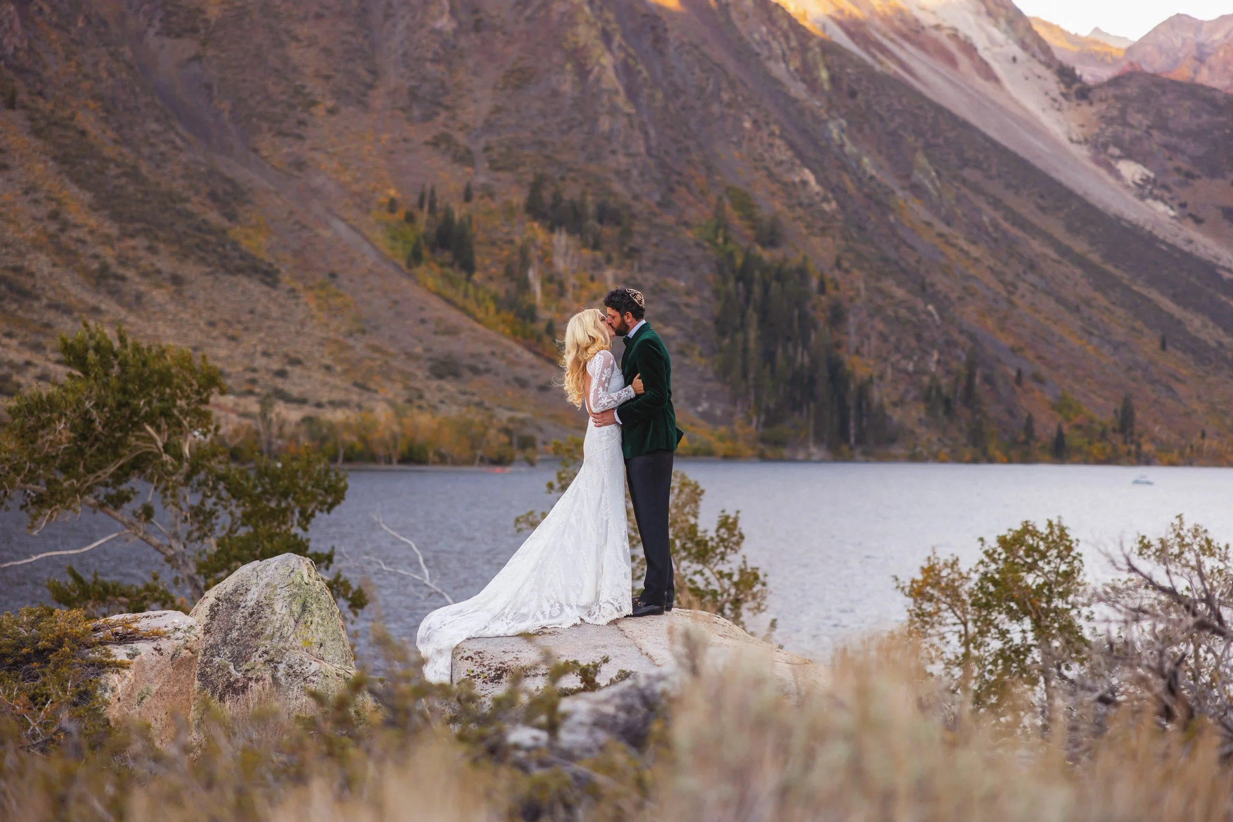A bride and groom in wedding attire embracing on a rock in front of a lake with mountains in the background.