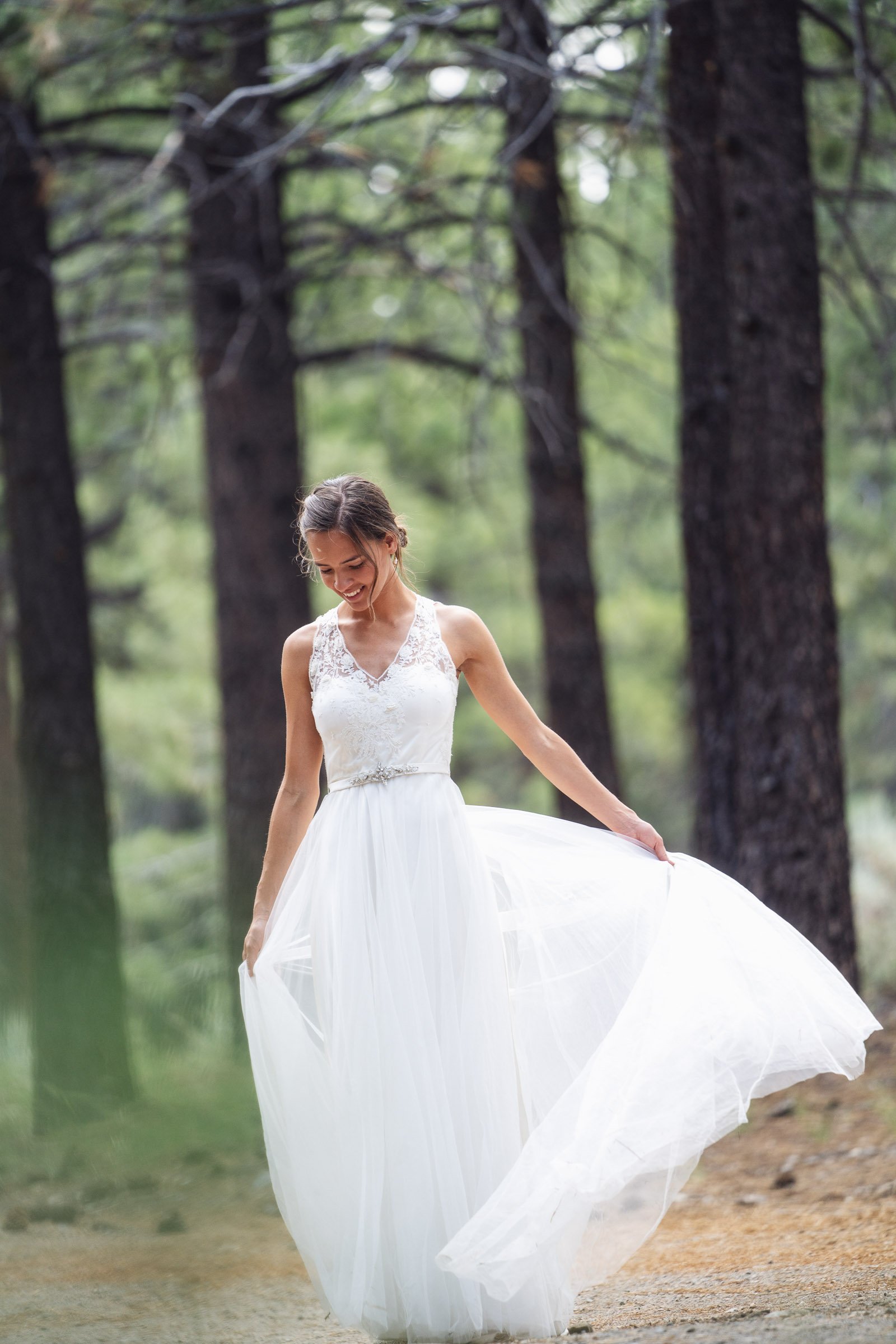 A woman in a white wedding dress walking through a forest, smiling and holding the edges of her dress.