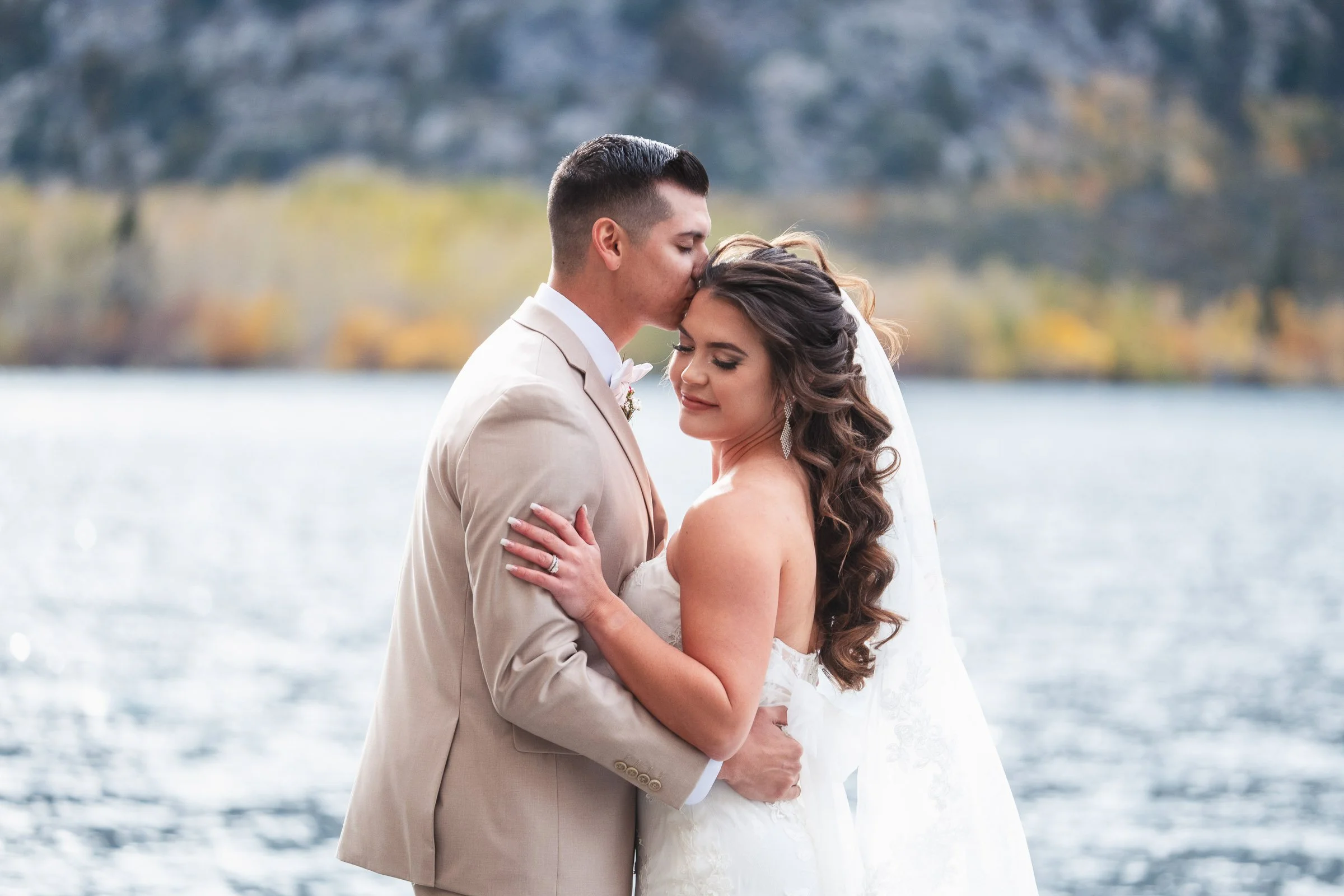 A bride and groom embracing by a body of water, with the groom kissing the bride's forehead and both smiling, in an outdoor wedding setting.