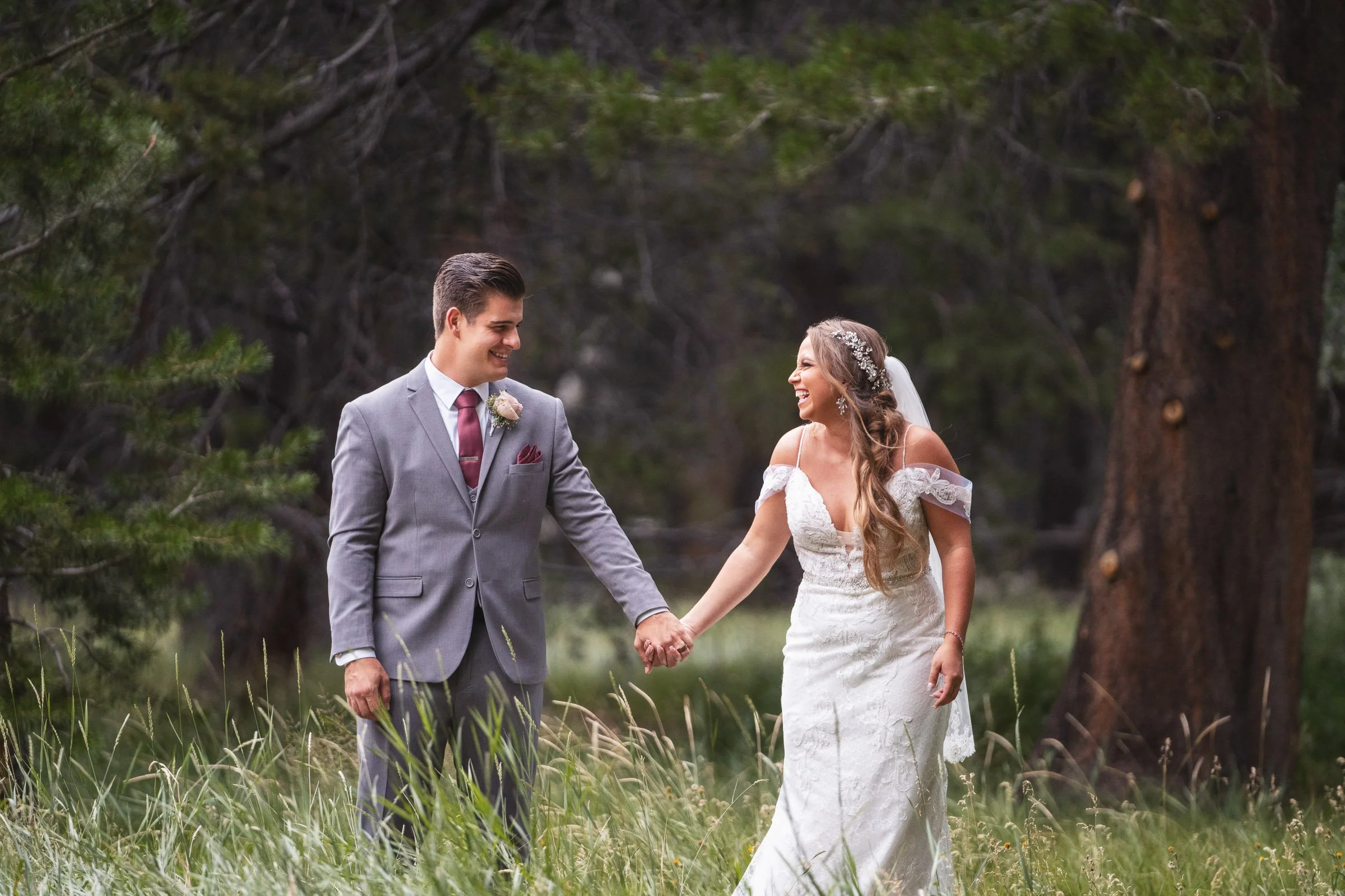 A bride and groom holding hands and smiling in a forested area during their wedding.