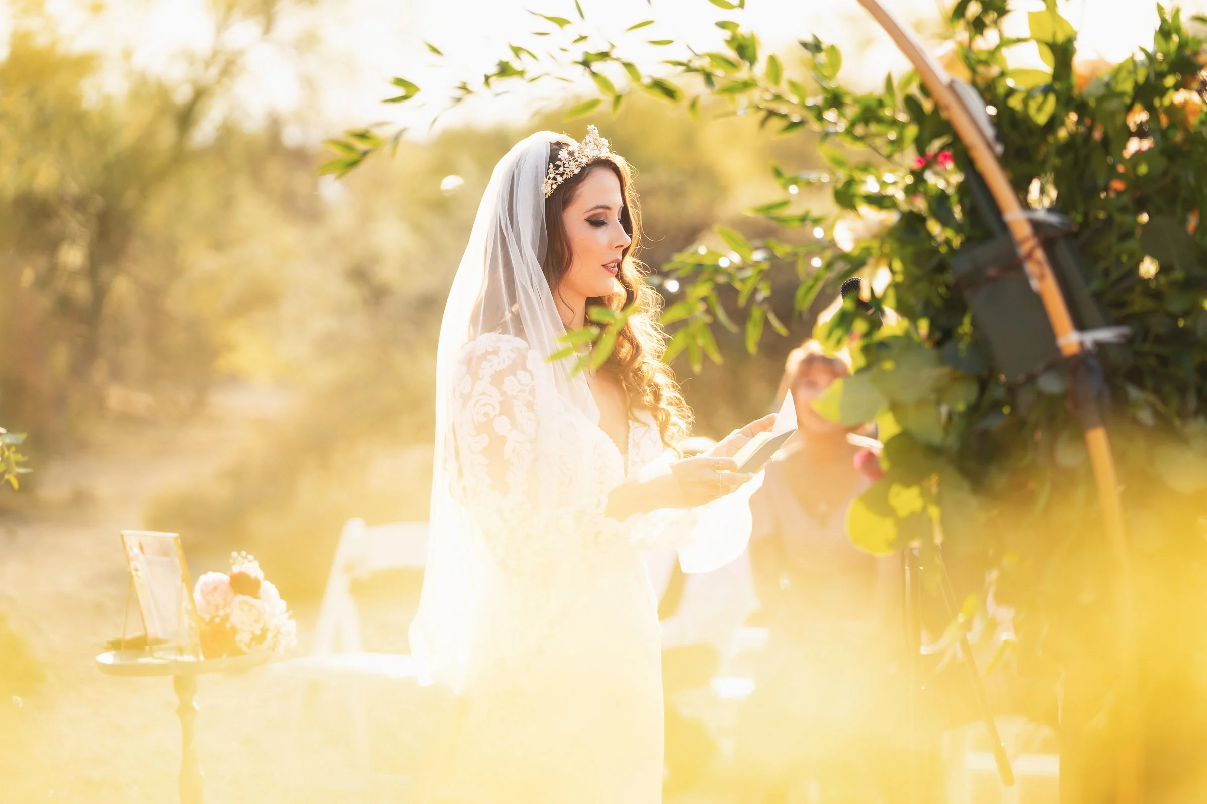 A bride with long wavy hair and a lace wedding dress, wearing a veil and floral headpiece, reading from a booklet outdoors during her wedding ceremony, with greenery and warm sunlight in the background.