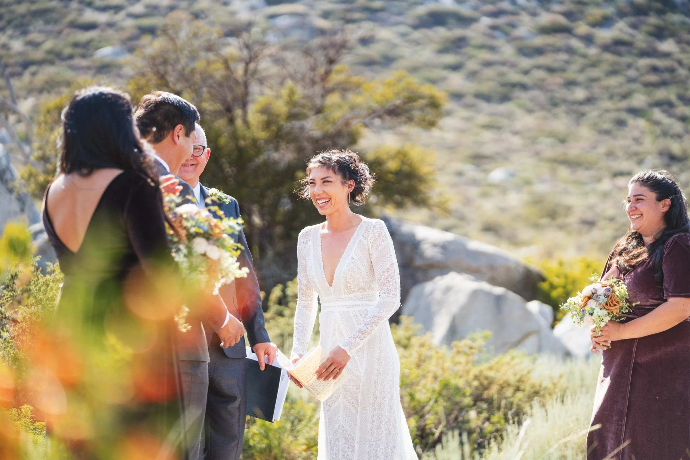 A bride in a white lace dress laughing during an outdoor wedding ceremony, with a groom and officiant on her left, and a bridesmaid in a purple dress holding a bouquet on her right, in a natural scenic setting with trees and rocks in the background.