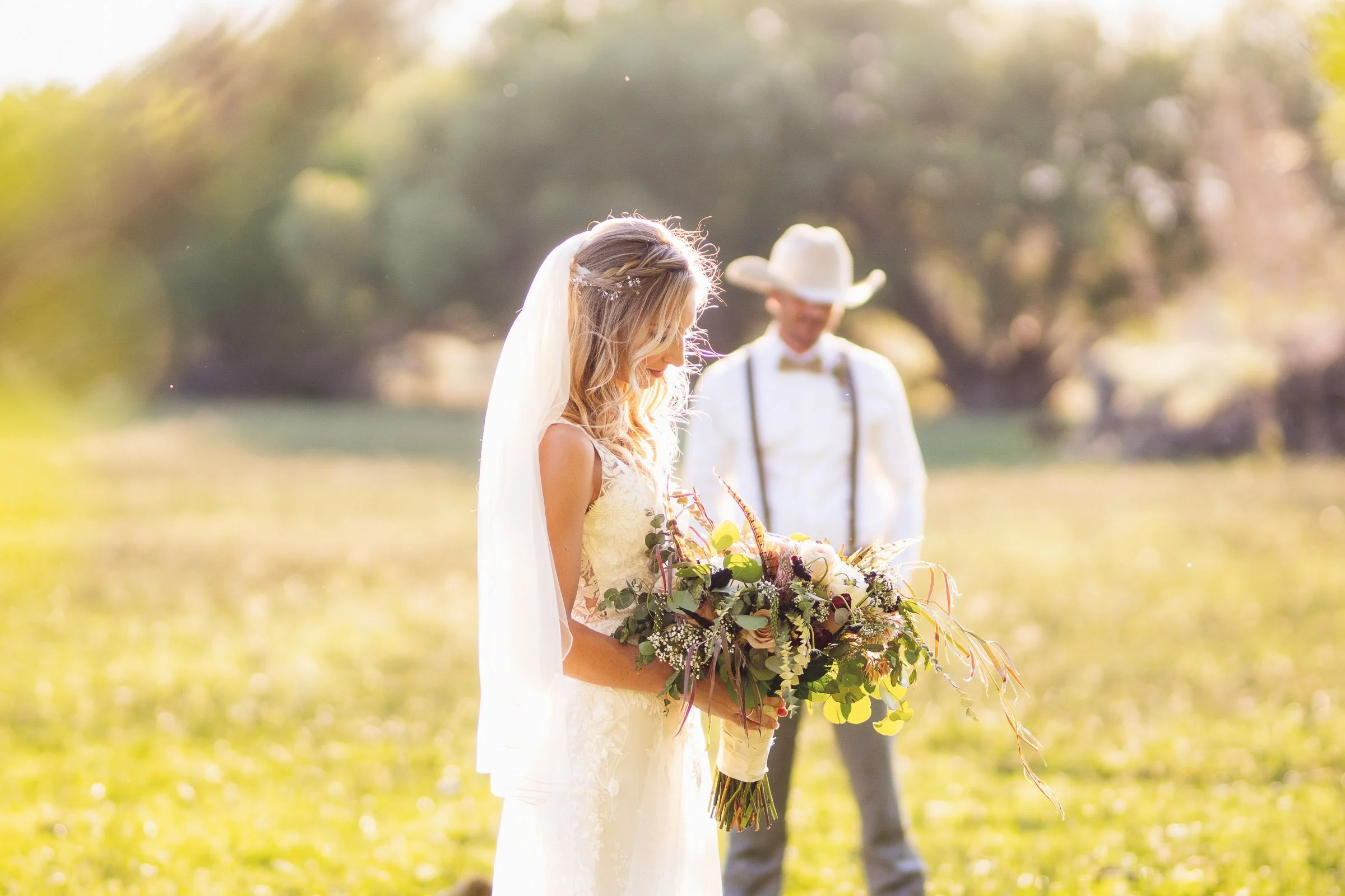 A bride in a white wedding dress holding a bouquet of flowers with a groom in a white shirt, suspenders, bow tie, and a cowboy hat standing behind her in an outdoor setting.