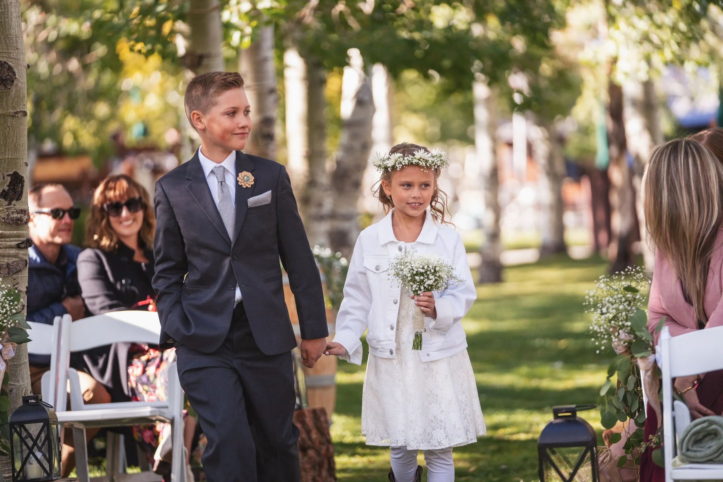 Young boy and girl holding hands during an outdoor wedding ceremony with guests seated nearby.