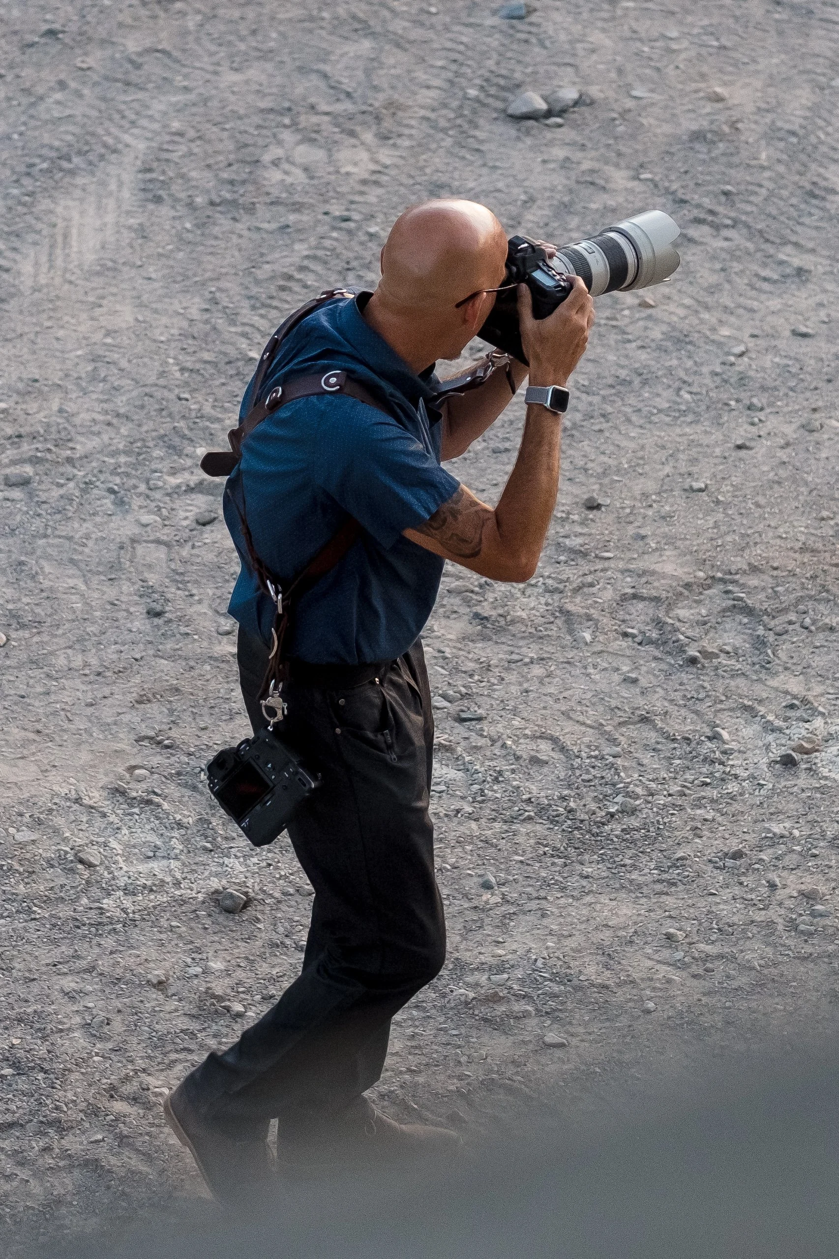 A bald man taking a photograph with a professional camera on a rocky outdoor surface.