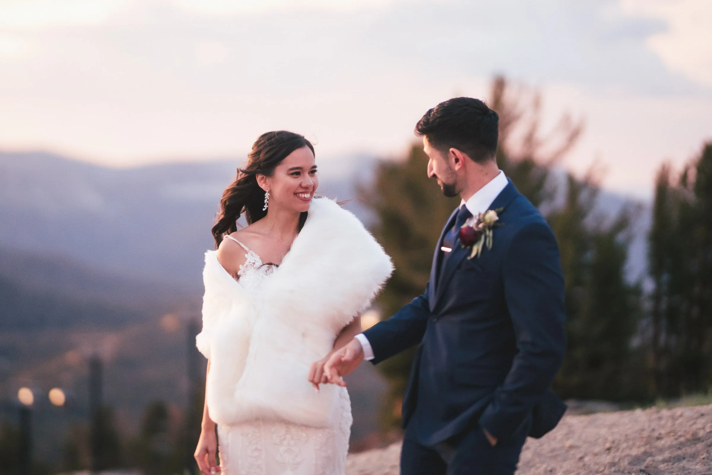 Bride and groom smiling at each other outdoors at sunset, holding hands, with mountains and trees in the background.