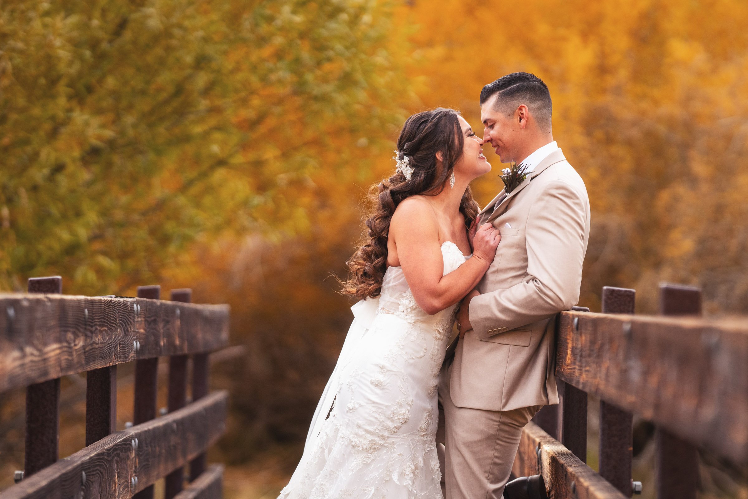 A bride and groom stand close together on a wooden bridge, smiling and about to kiss, with autumn leaves in the background.
