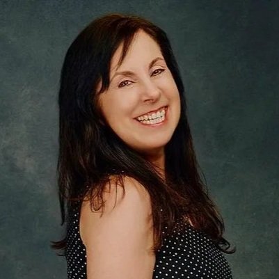 A woman with dark hair smiling and wearing a black polka dot top against a dark background.