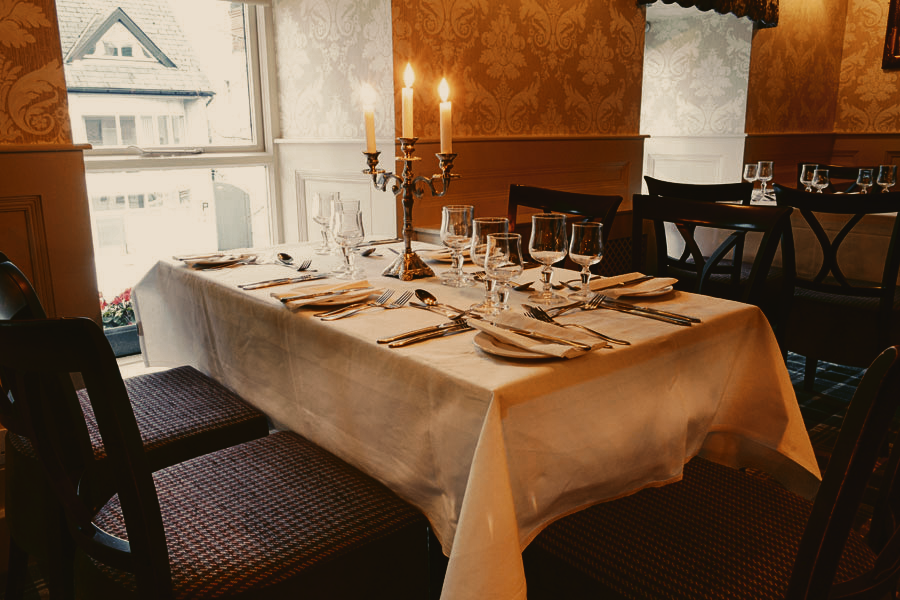 Elegantly set dining table with a white tablecloth, multiple wine glasses, silverware, and folded napkins, decorated with a candelabrum holding lit candles, in a cozy, warmly lit restaurant with patterned wallpaper and large windows showing a snowy scene outside.