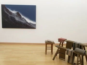 Leinwand mit Berglandschaft hängt an einer weißen Wand, im Vordergrund befinden sich mehrere gestapelte Teppiche auf Klappbferen.