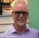 A smiling man with glasses and a checkered shirt, standing indoors with mint green walls and a framed picture in the background.