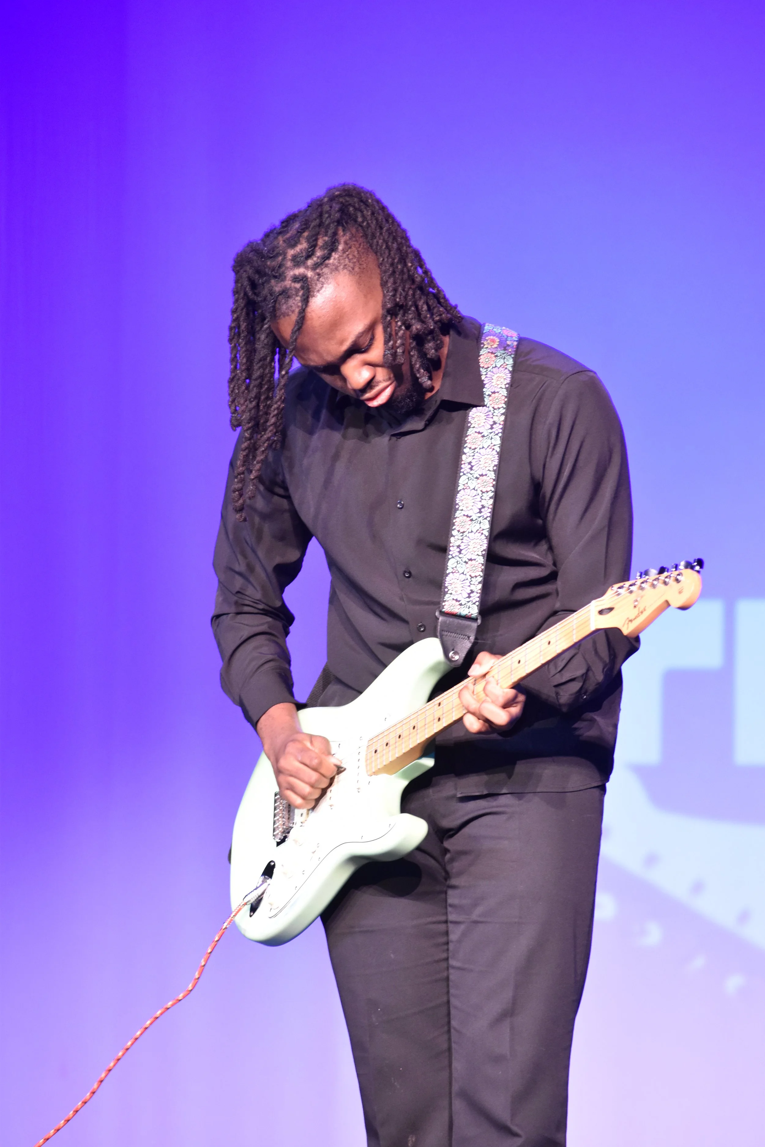 A man playing a white electric guitar on stage with a purple background.