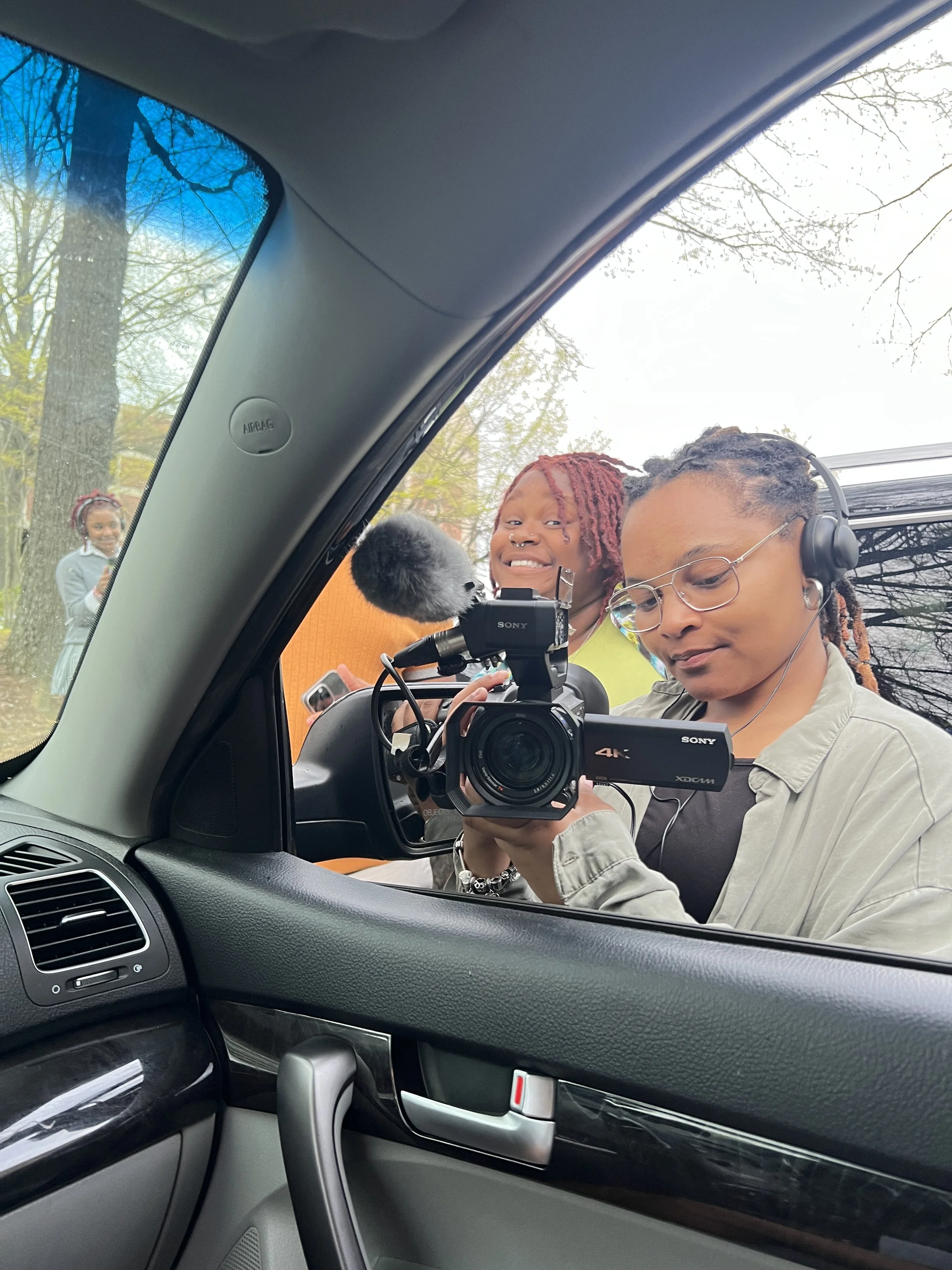 Two women filming a video with a professional camera from inside a car, with a person standing outside in the background.