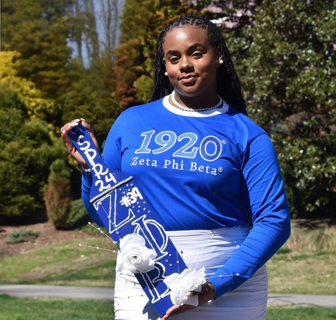Woman in a blue shirt with white lettering holding blue and white decorated graduation sash with white flowers, outdoors with trees in the background.