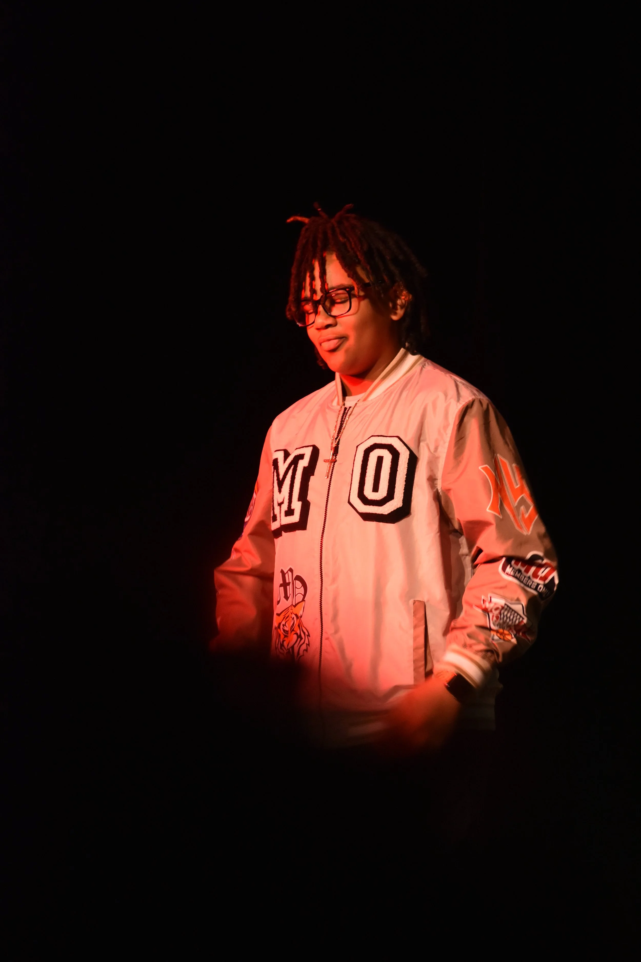 A person with glasses and dreadlocks wearing a beige jacket with various patches, standing against a black background, illuminated by red light.