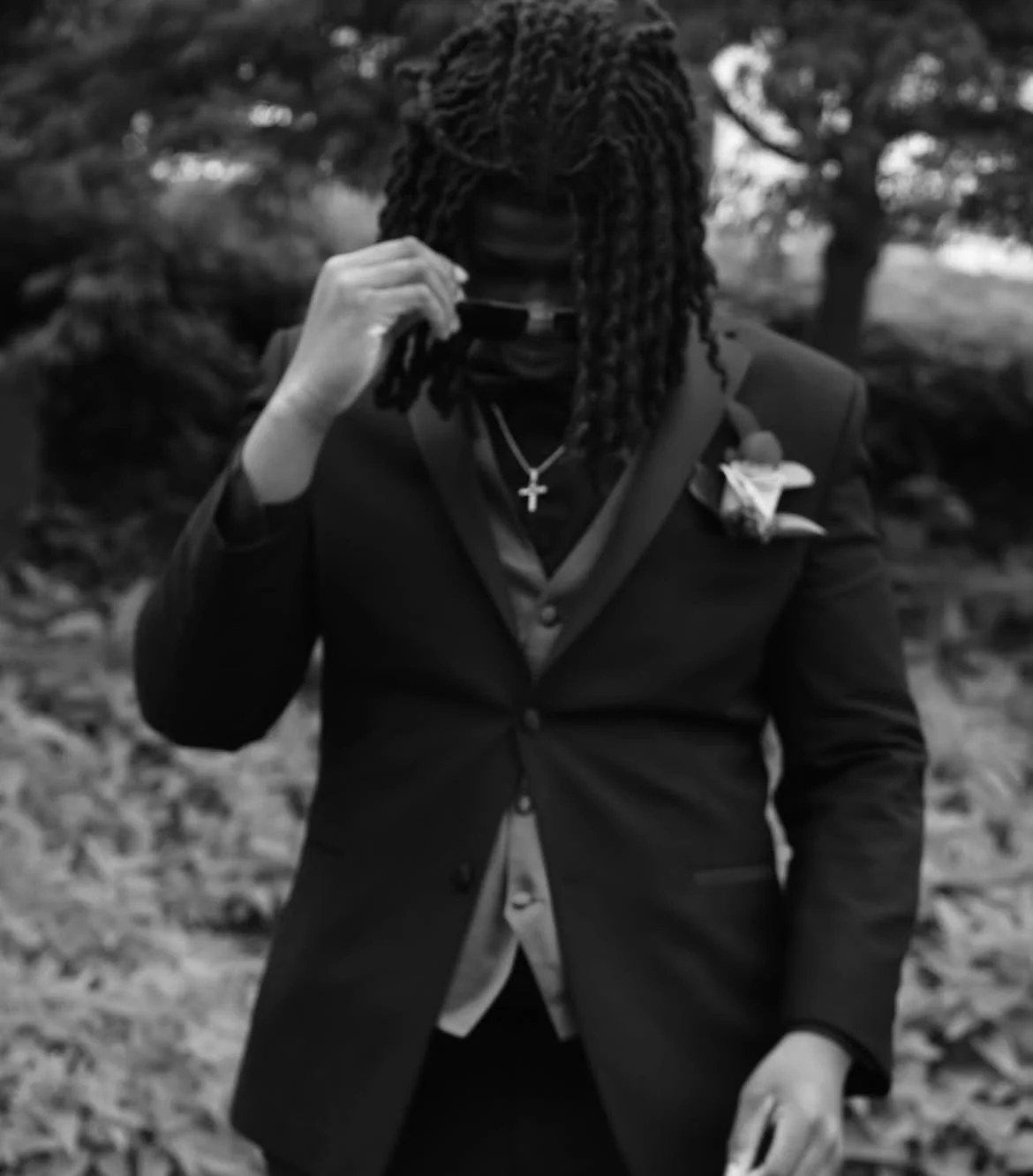 A man with long dreadlocks wearing a dark blazer, shirt, and cross necklace outdoors, holding sunglasses near his face with a blurred background.