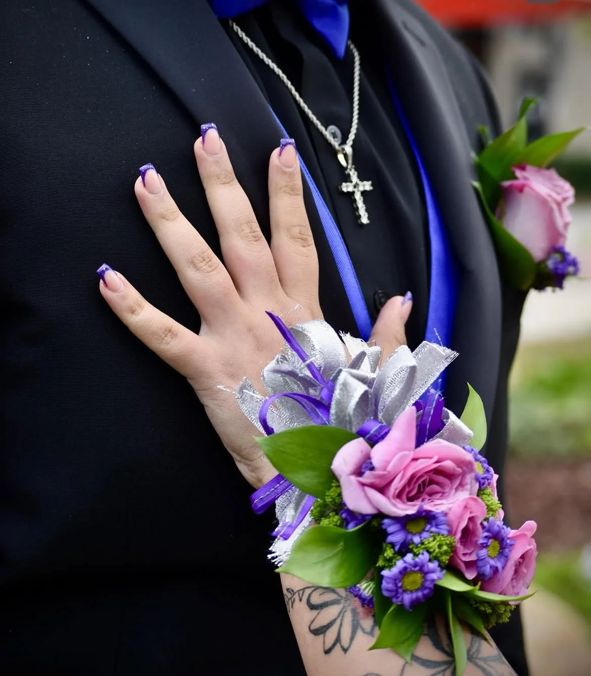 Close-up of a woman's hand with purple and silver nails, wearing a corsage with pink roses, purple flowers, and green leaves, placed on a man's chest with a black suit and a silver cross necklace, during a wedding or formal event.