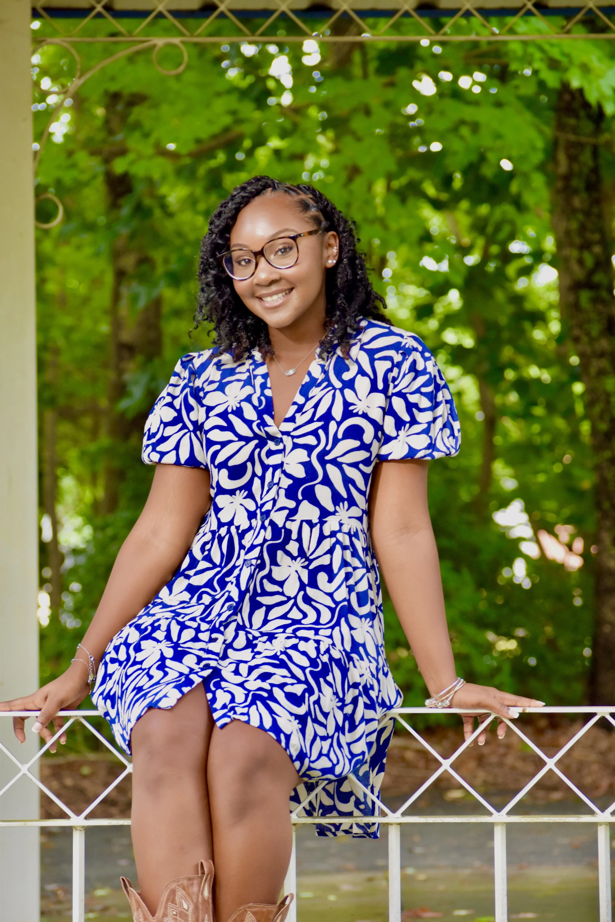 A young woman sitting outdoors on a white picket fence, wearing a blue and white floral dress and cowboy boots, with a background of green trees.