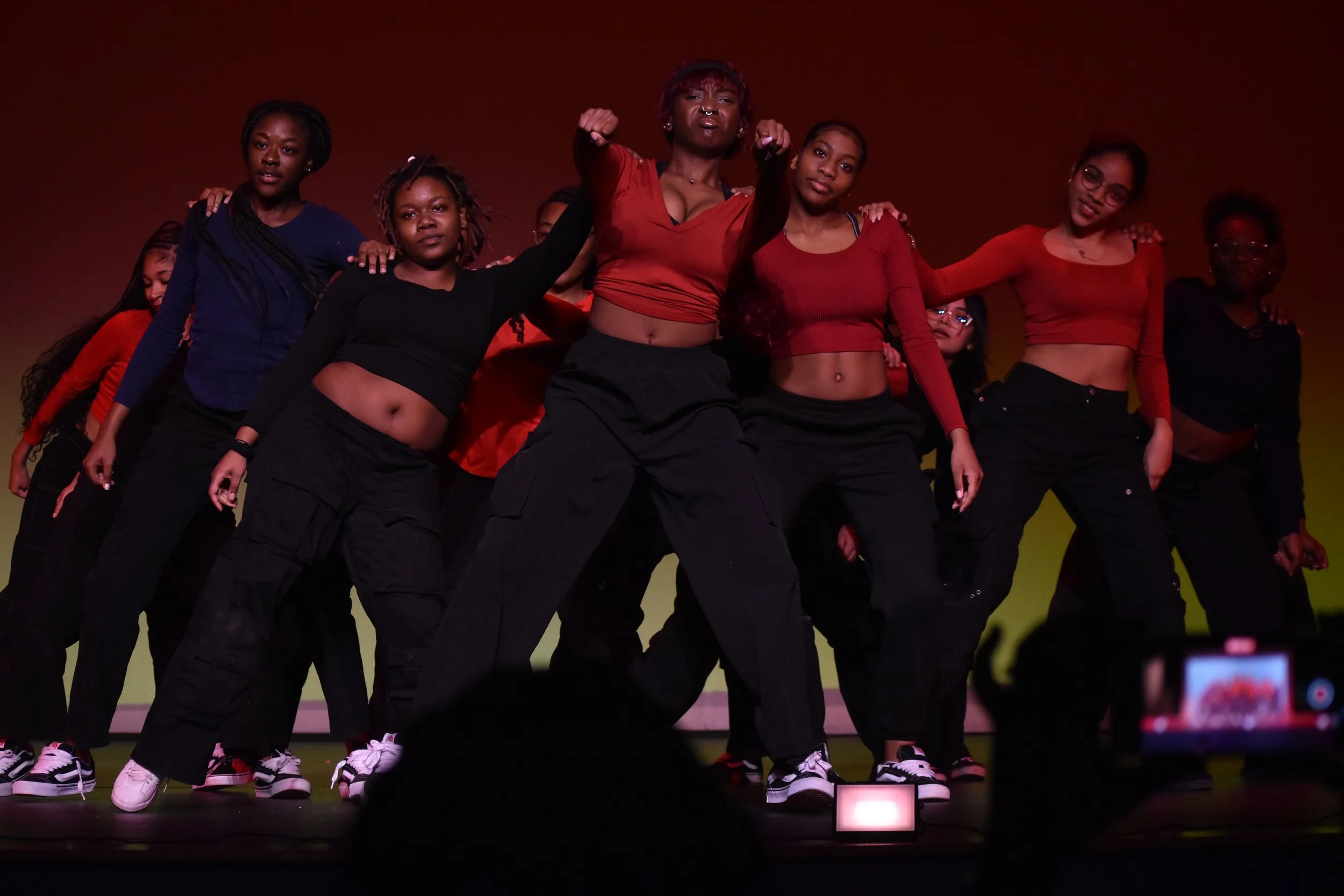 Group of young women performing a dance on stage, wearing casual black and red outfits, with a reddish backdrop.