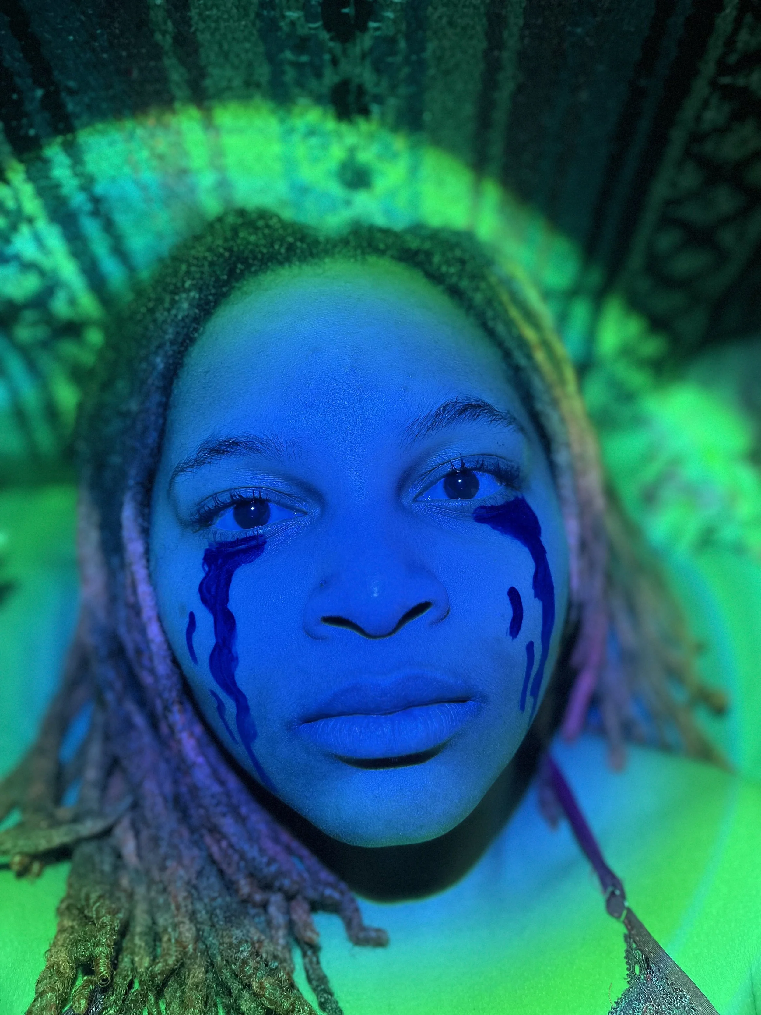 A person with dreadlocks wearing bright makeup and face paint, illuminated by vibrant green and blue lighting.