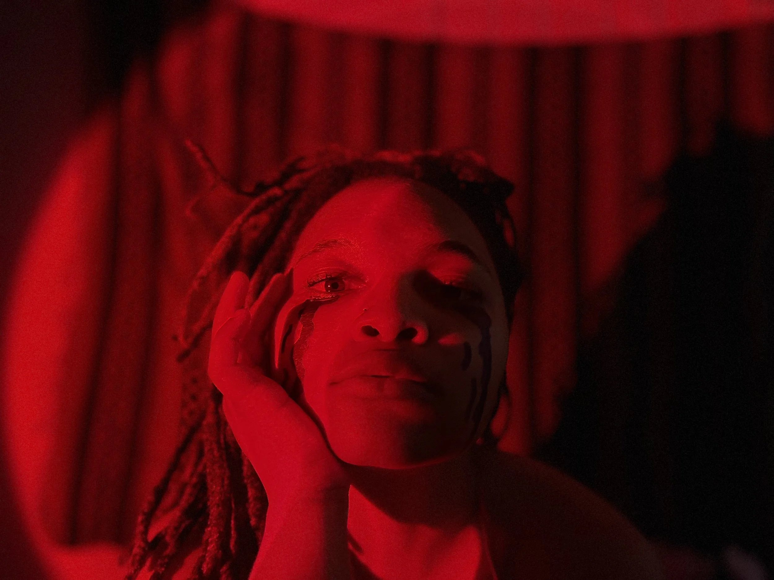 A woman with dreadlocks and face paint, lying down in red lighting with a curtain background.