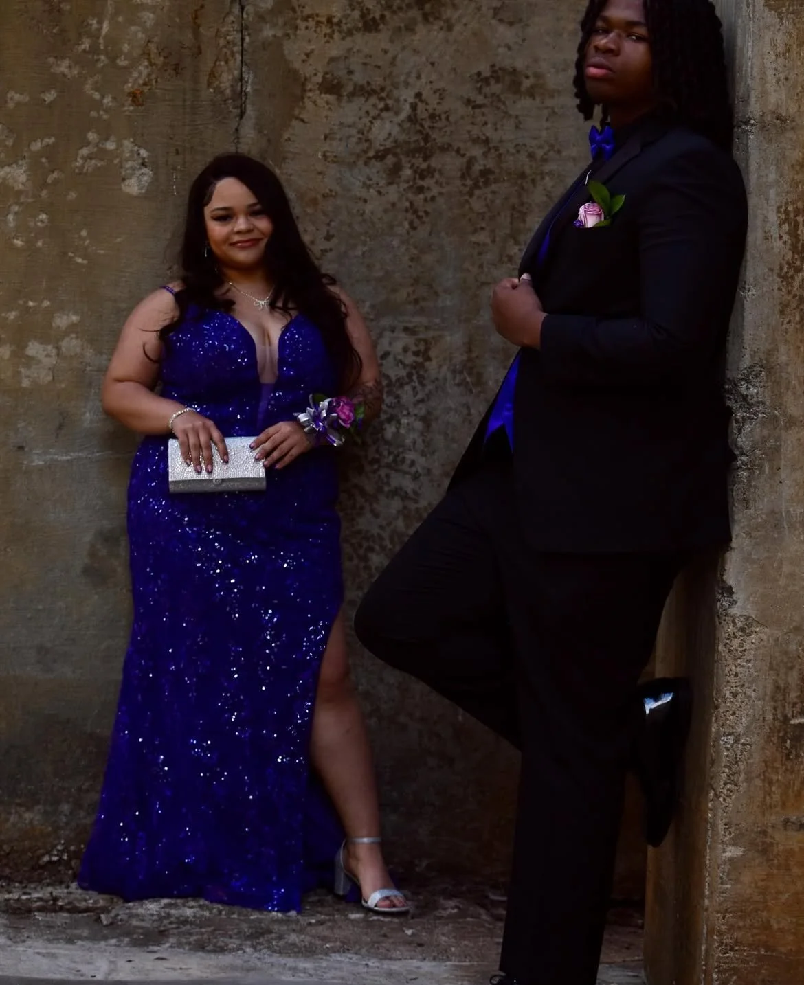 Two young adults dressed in formal attire, posing against a textured stone wall. The woman is wearing a sparkling blue gown and holding a silver clutch, while the man is dressed in a black suit with a blue shirt and bow tie, with boutonniere.